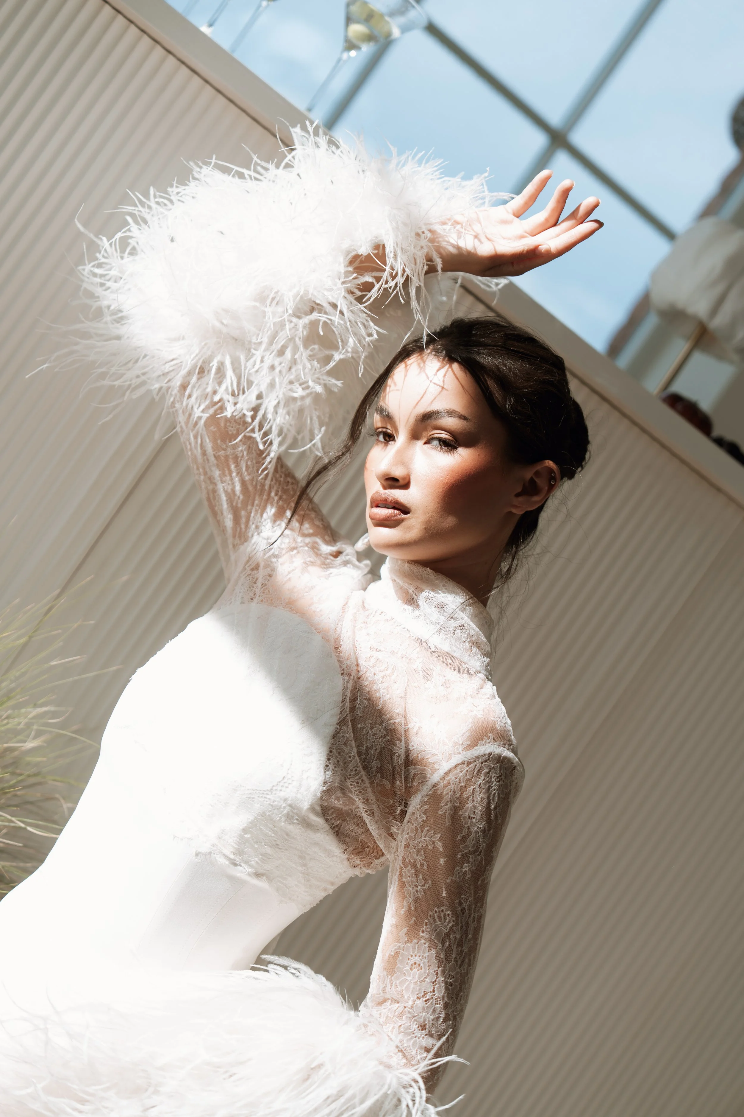 A woman in a white lace dress with feathered sleeves posing indoors against a background of a glass ceiling and light-colored wall, looking at the camera with an intense expression.