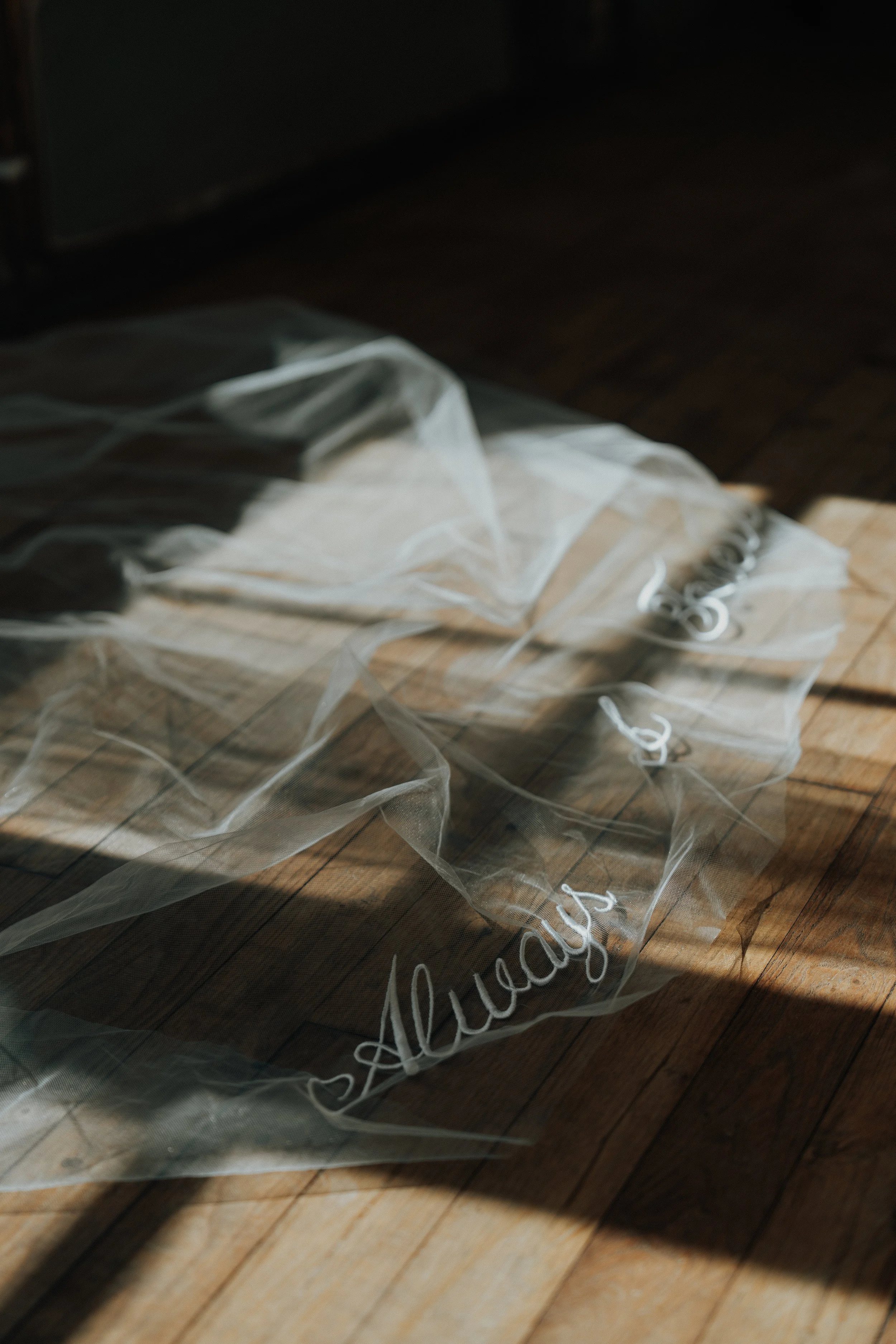An editorial, contemporary bridal detail shot of a sheer wedding veil laid out on a wooden floor, featuring custom white cursive calligraphy that reads "now and forever and always," beautifully illuminated by artistic natural sunlight and shadows.