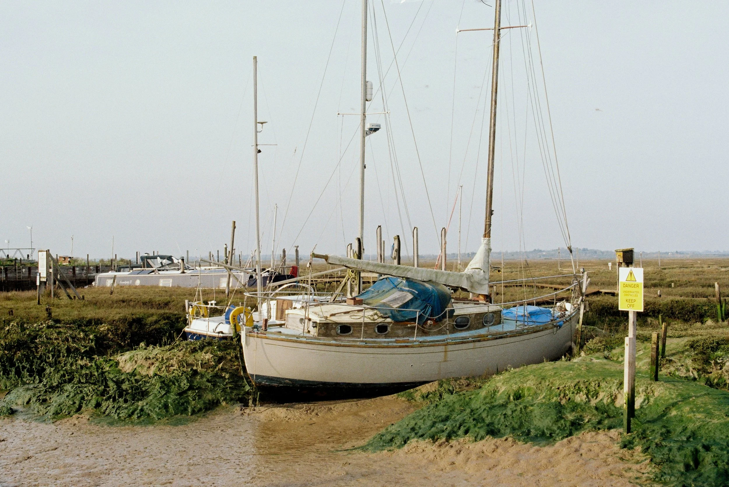 A boat is stranded on a muddy riverbank with grass and weeds, surrounded by a fence and yellow warning signs, under a cloudy sky.