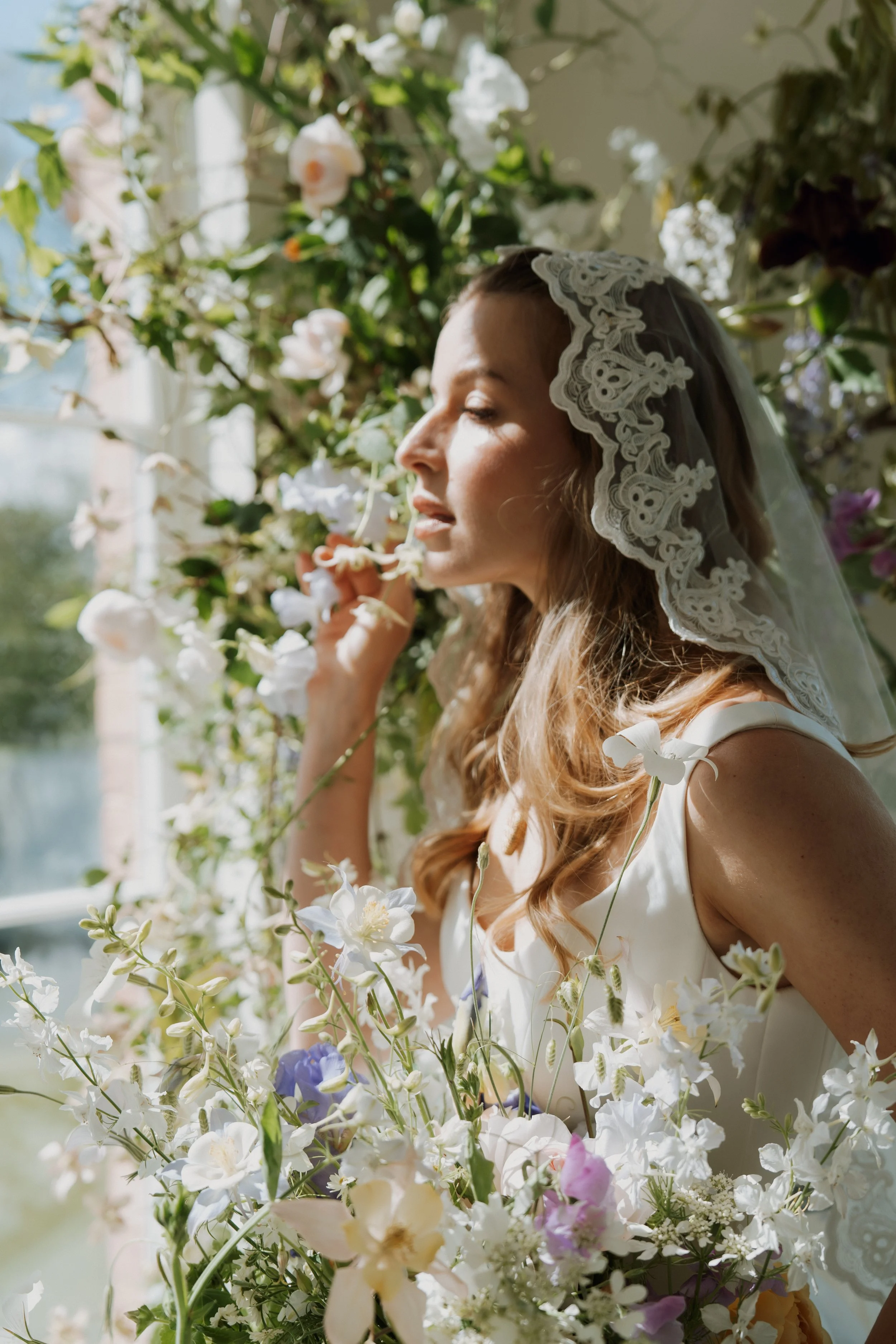 An elegant bride with long, wavy hair wearing a Savannah Miller Bridal wedding dress and lace veil, holding a bouquet of white and pastel flowers against a lush backdrop of greenery.