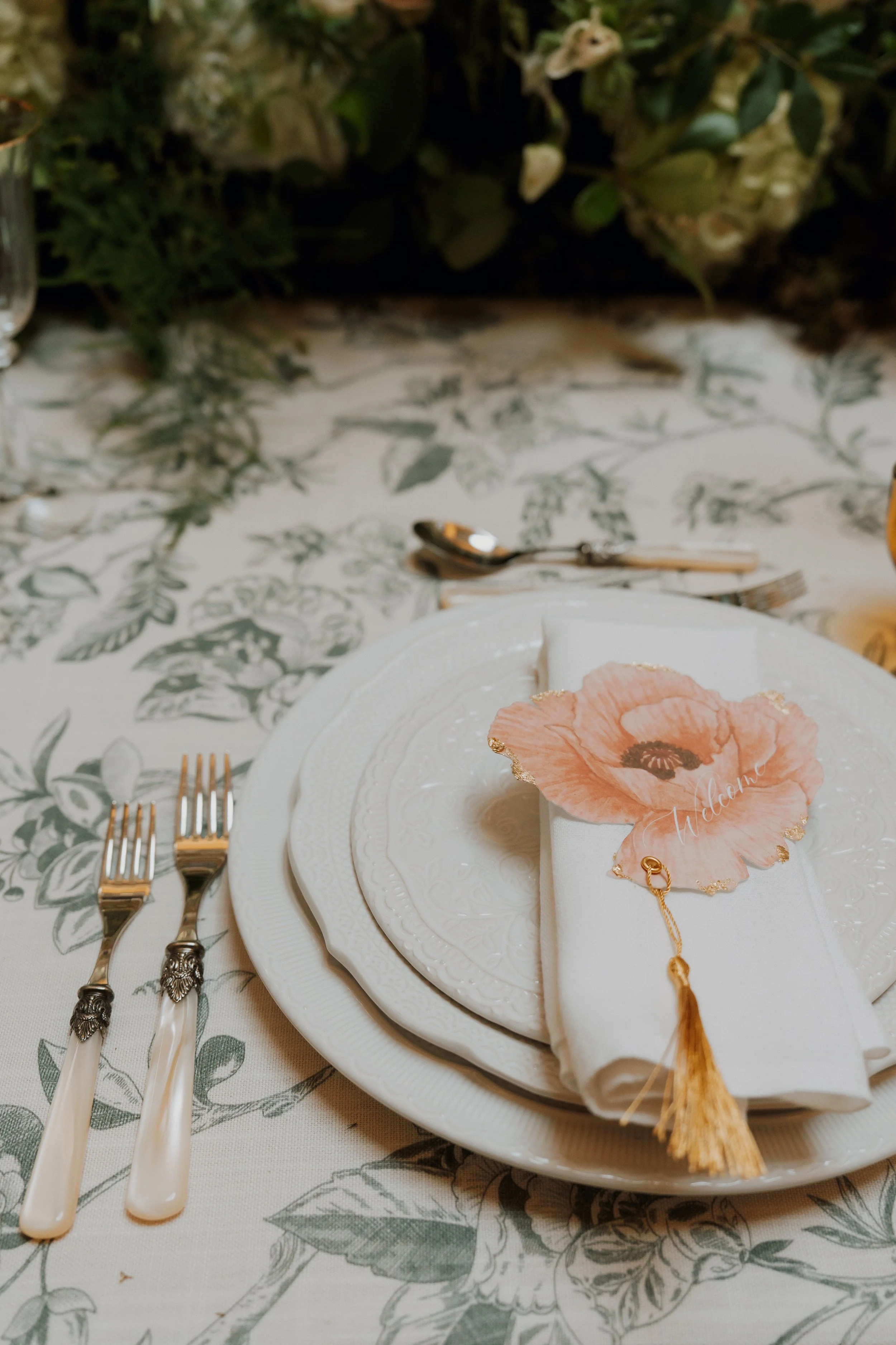 An elegant luxury tablescape featuring white plates, polished gold utensils, and a bespoke pink floral 'Welcome' menu card with a gold tassel on a patterned tablecloth.