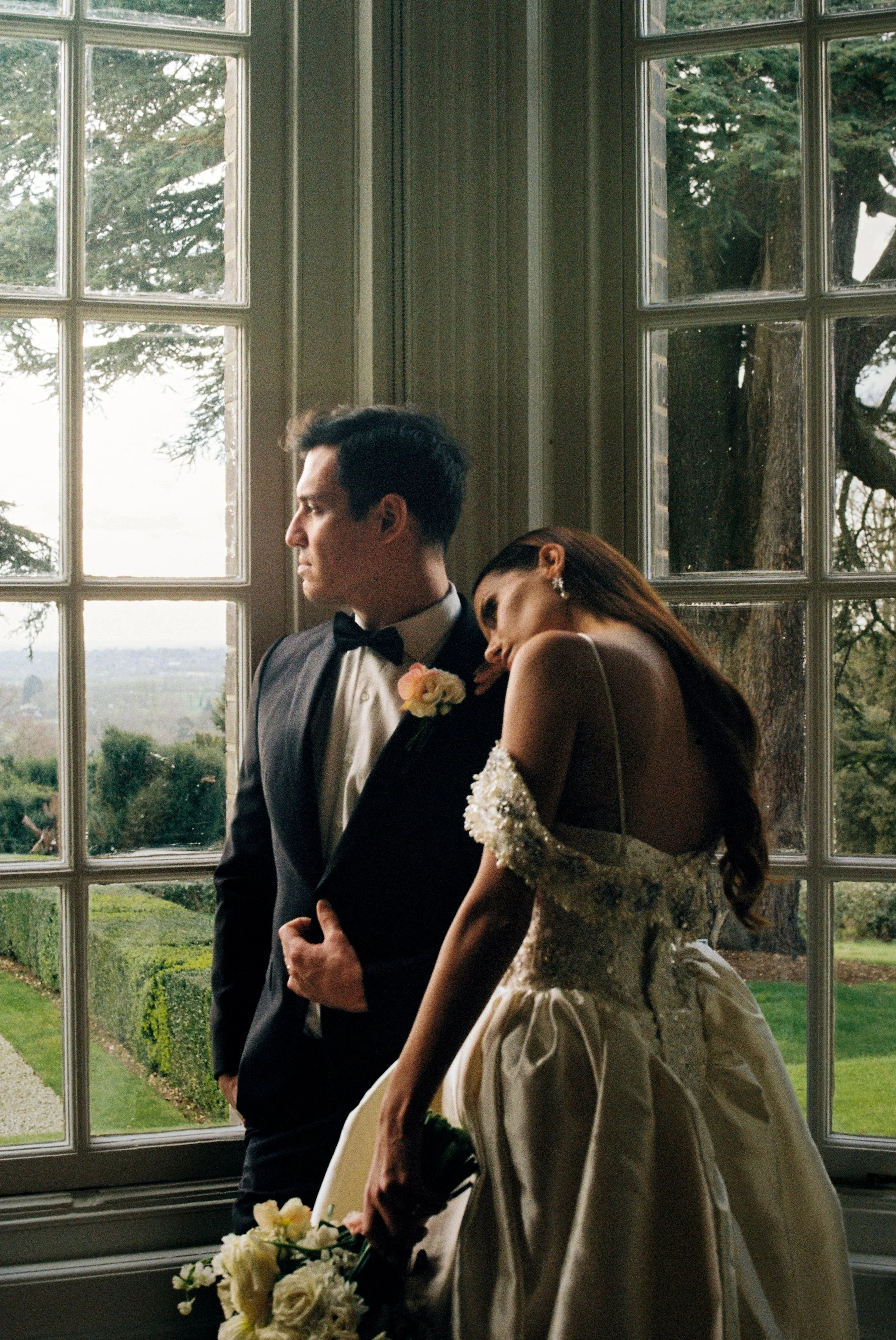A bride and groom standing near large window with a view of green trees and fields outside. The groom is dressed in a black tuxedo with a bow tie, and the bride is wearing a gown with floral details on the sleeve, holding a bouquet of white flowers.