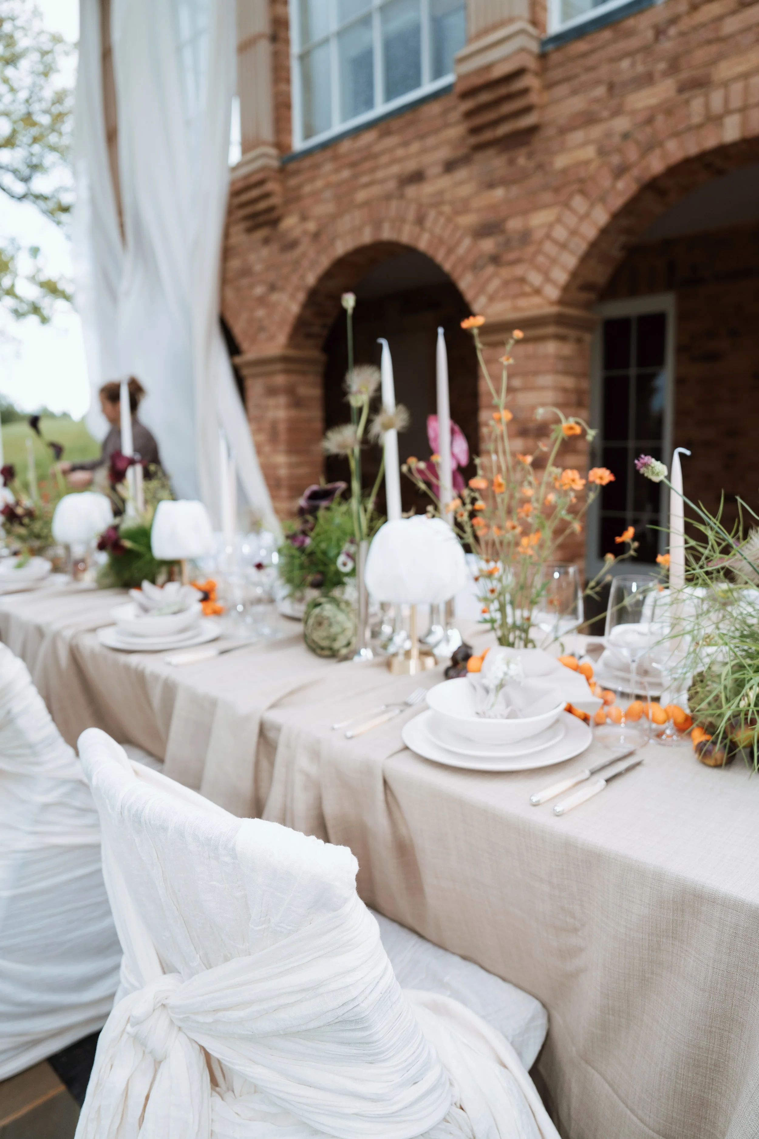 An elegant outdoor wedding tablescape featuring a long dining table beautifully decorated with luxury candle holders, white flowers, and fine tableware, set against a historic brick building.