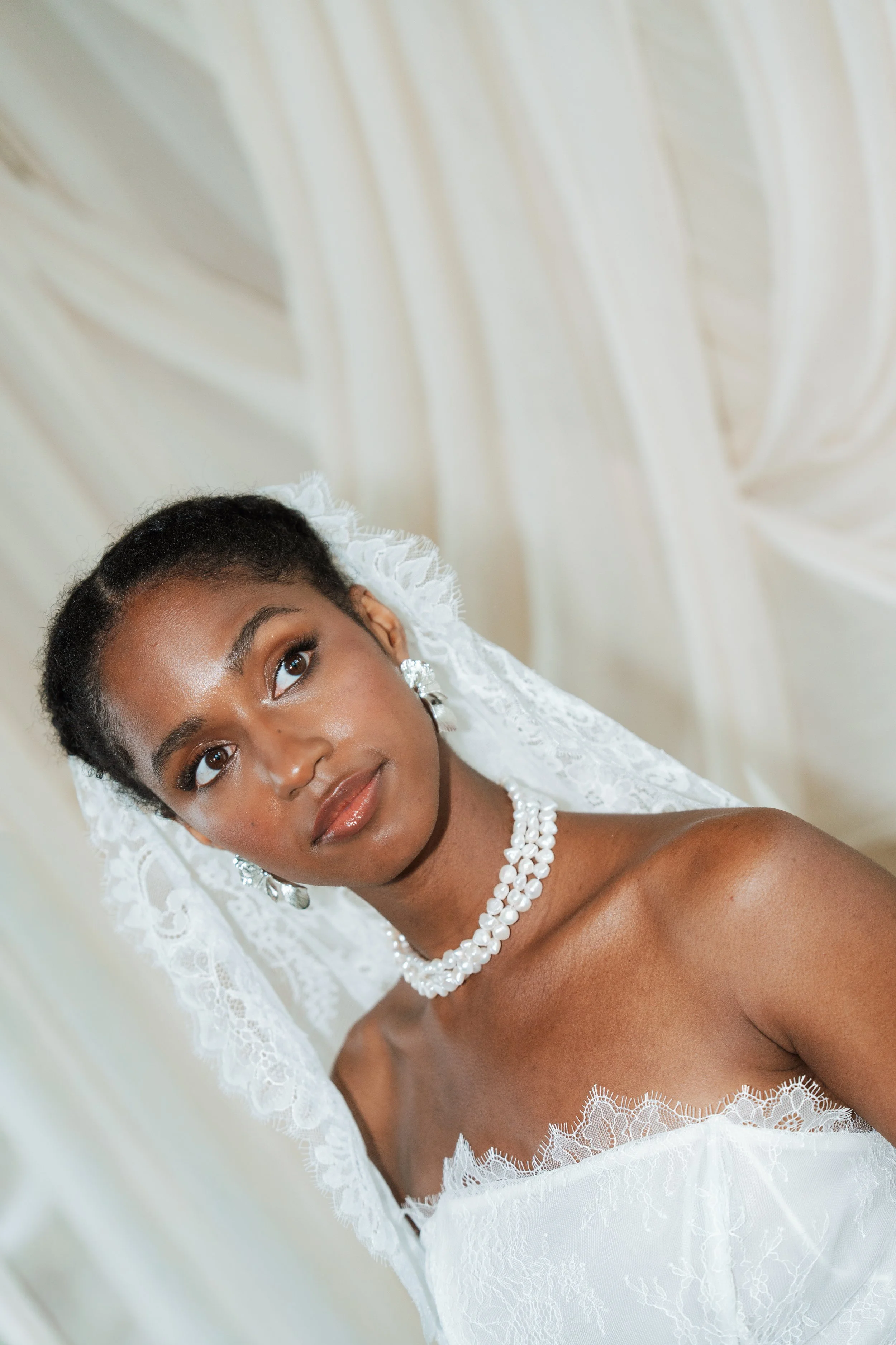 Close-up portrait of an elegant Black bride wearing a white lace bridal veil and pearl jewellery.