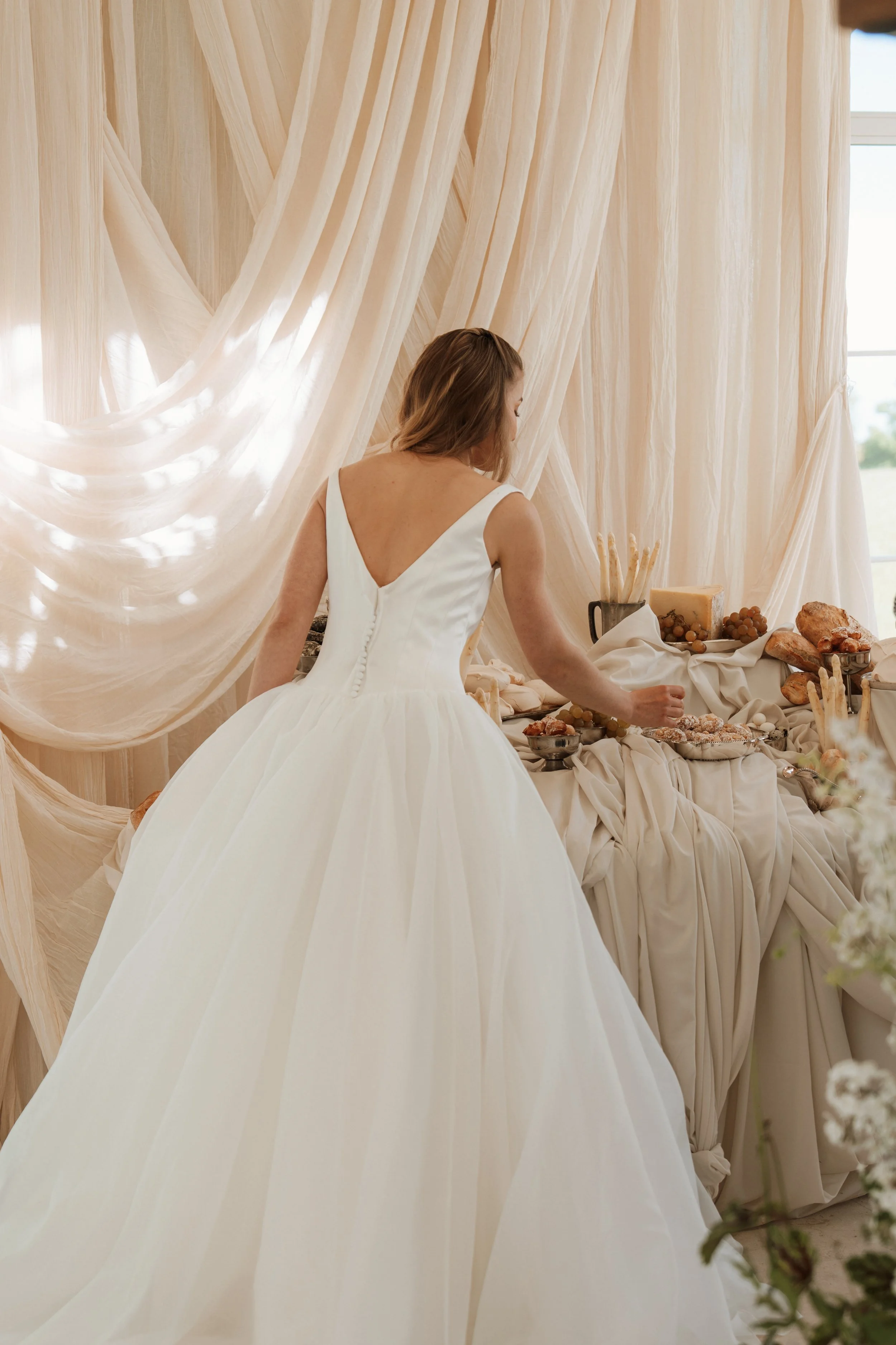 Rear view of a bride standing in an elegant Savannah Miller Bridal wedding dress next to a luxury grazing table with artisan bread and cheese, set against cream curtains.