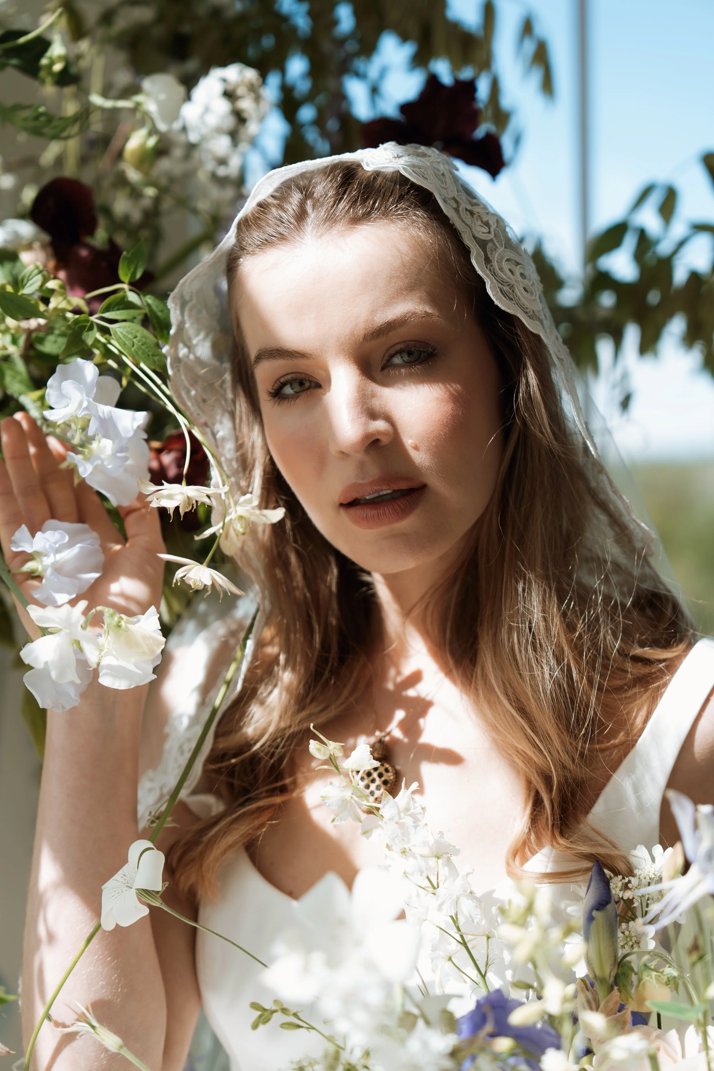 Close-up bridal portrait of a woman with blue eyes wearing a Savannah Miller Bridal wedding dress and lace veil, surrounded by elegant white and purple flowers.