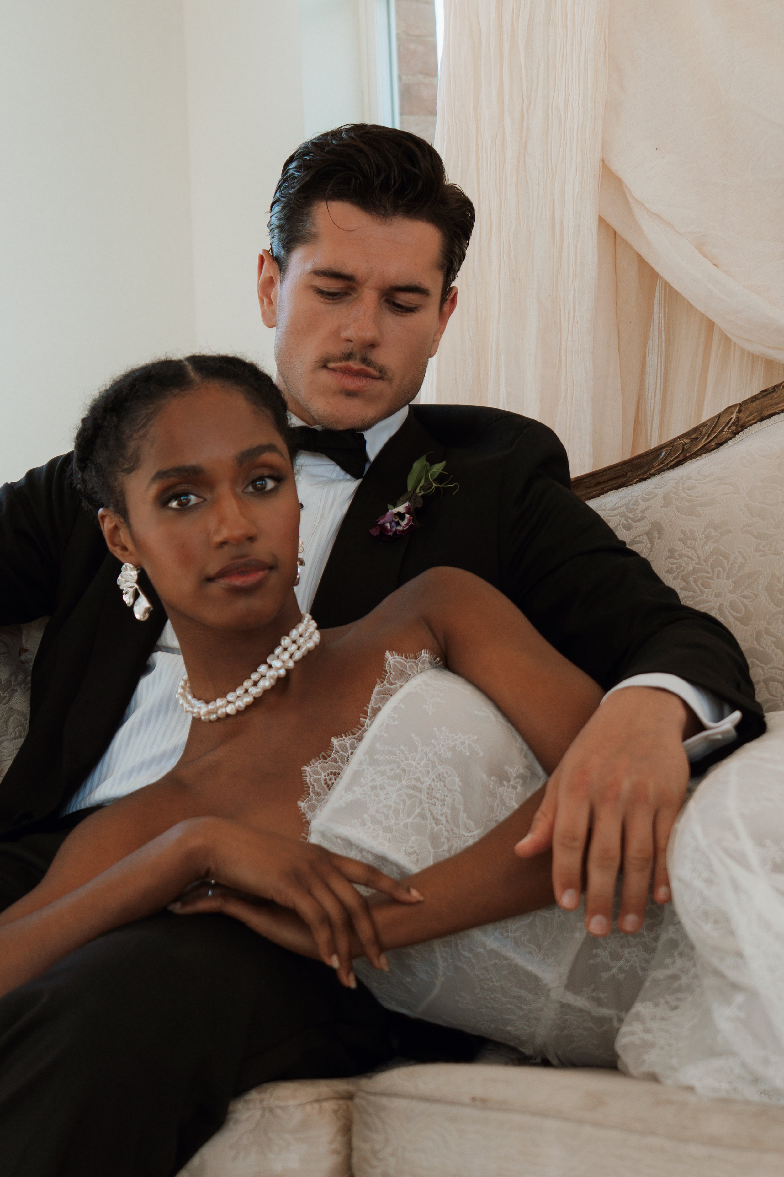 An intimate portrait of a newlywed couple sitting closely in a softly lit room; the bride wears an elegant Vera Wang wedding dress with pearl jewelry, alongside the groom in a classic tuxedo.