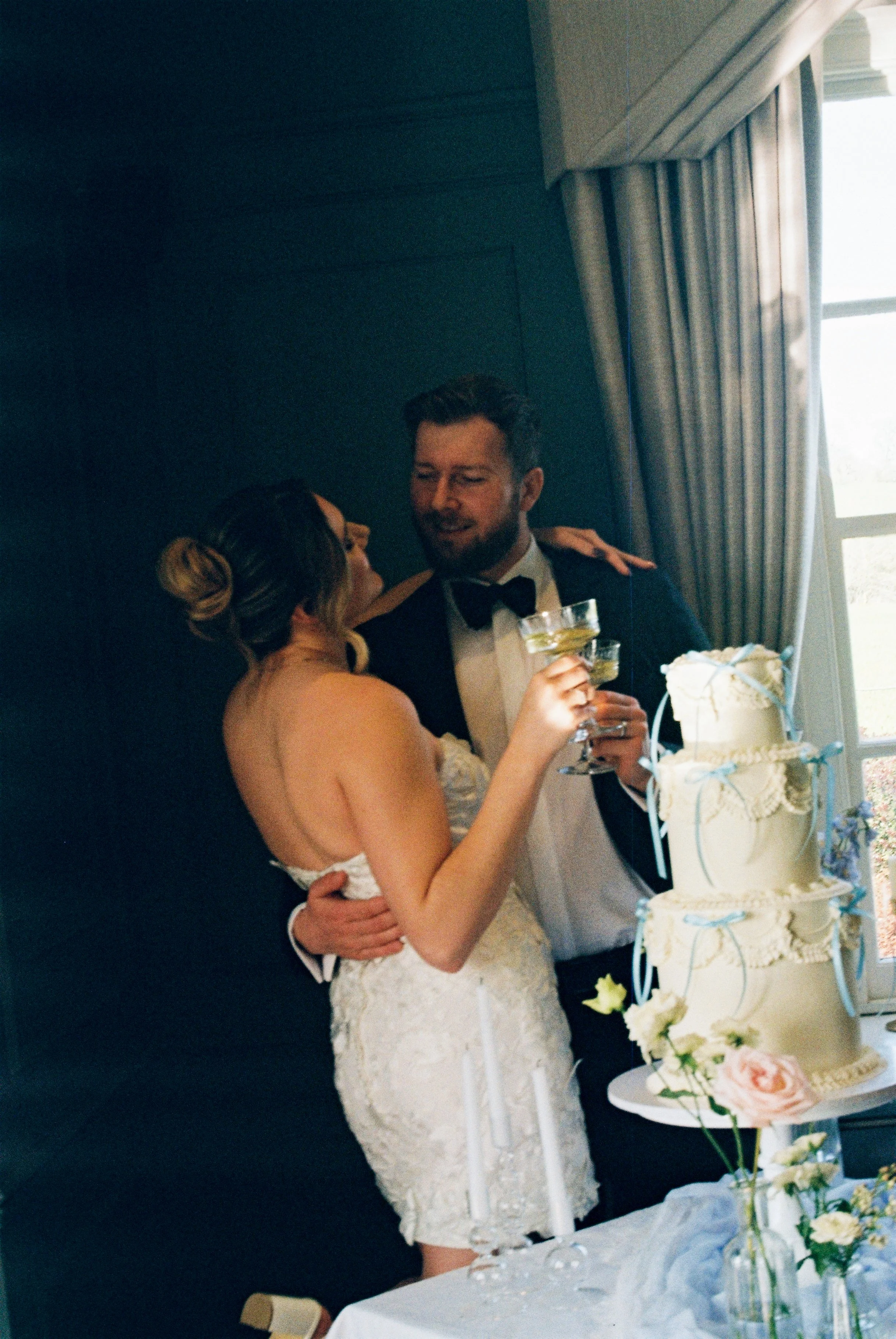 A bride and groom stand close together, smiling and holding glasses at their wedding reception. There is a tiered wedding cake decorated with flowers on the table in front of them. The groom wears a tuxedo with a bow tie, and the bride has her hair s