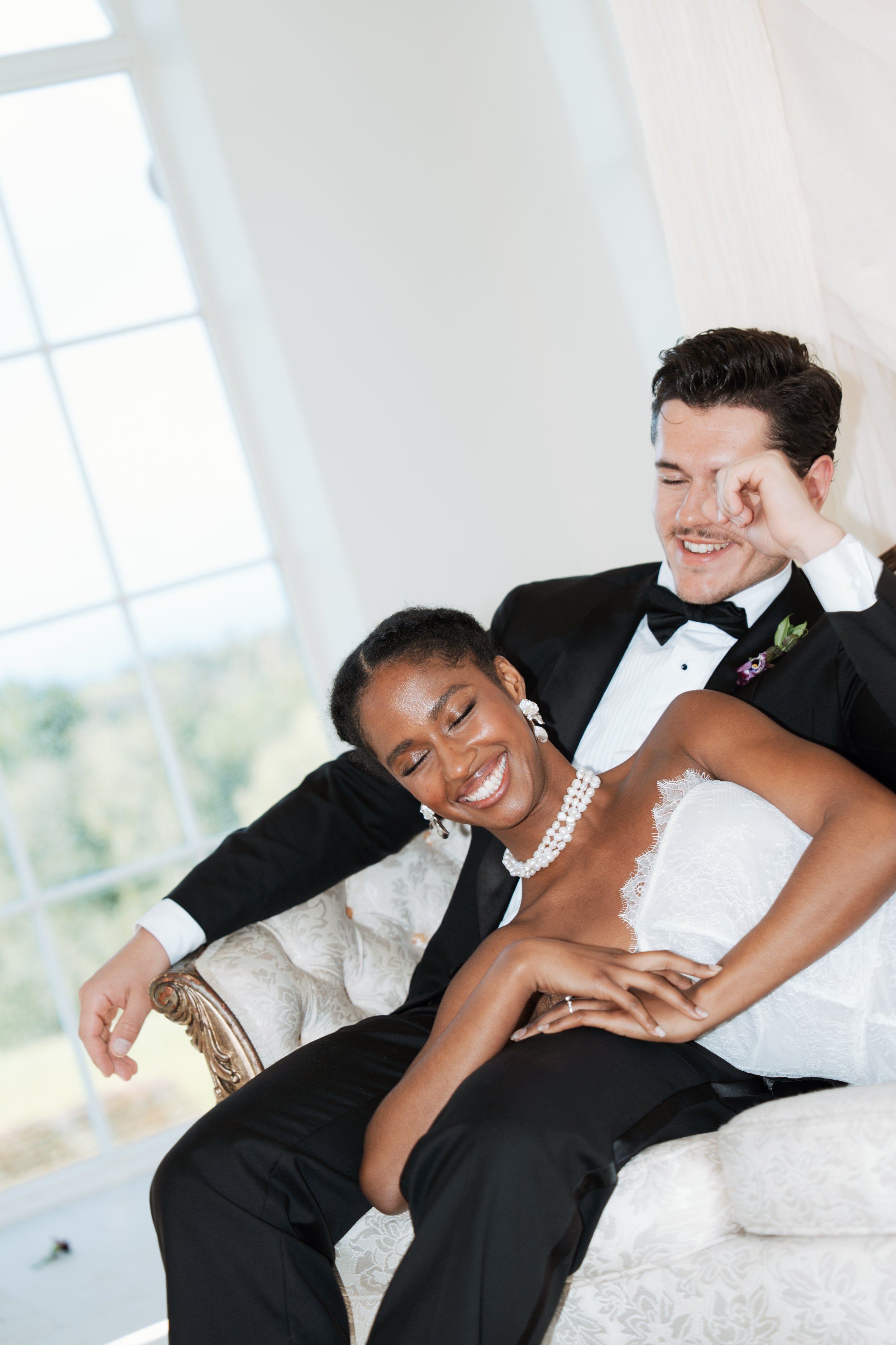 A candid, joyful portrait of a newly married couple laughing and sharing a happy moment together on a vintage sofa in front of large, sunlit windows.
