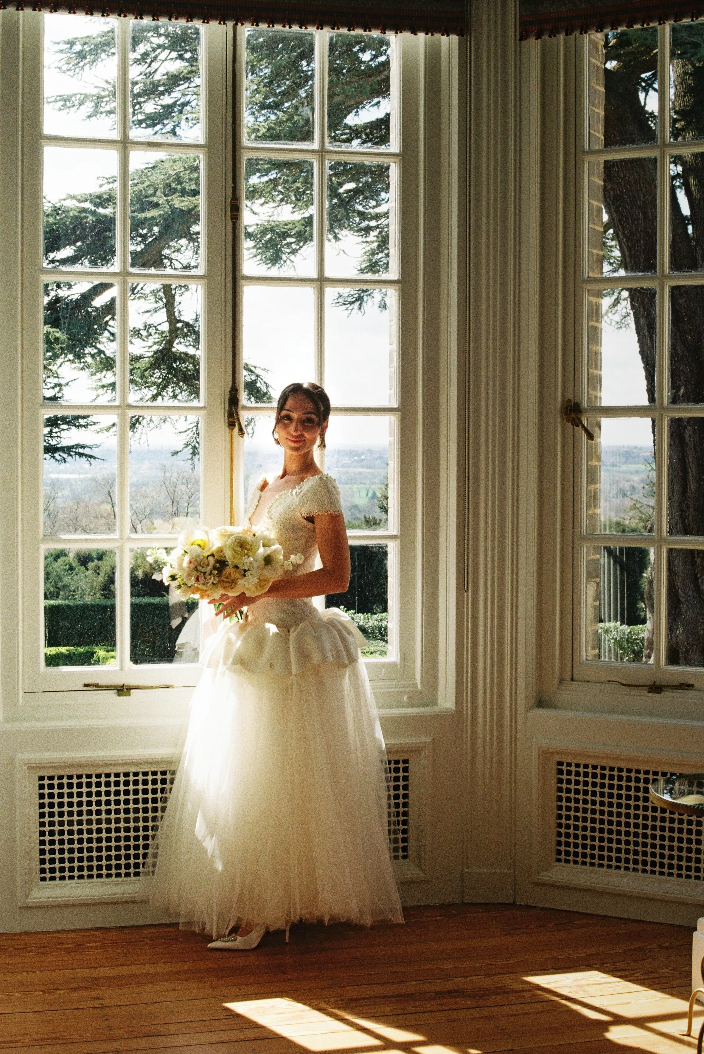 A bride standing in front of large windows with trees outside, wearing a white wedding dress and holding a bouquet of flowers.