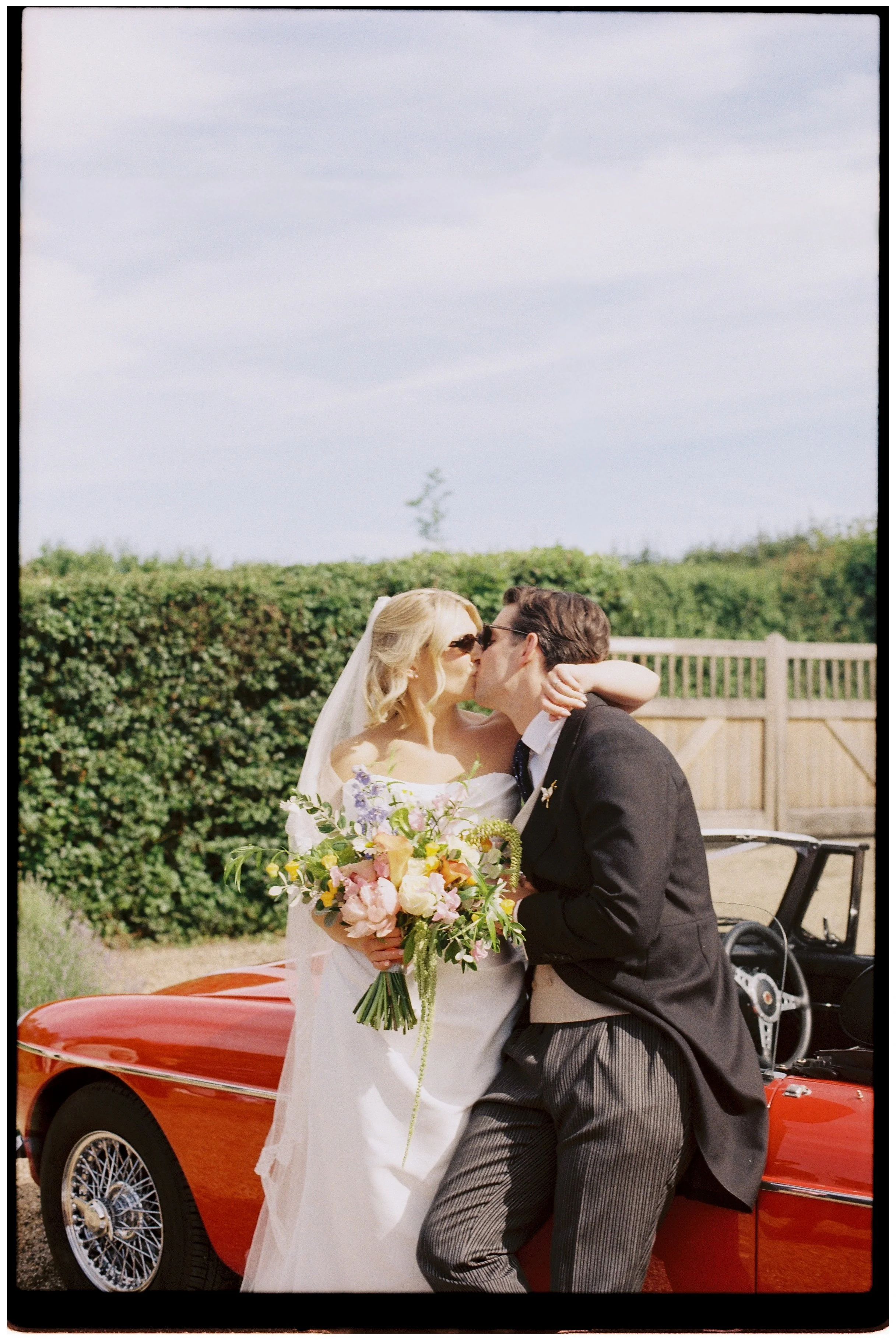 Bride and groom kissing beside a vintage red MG at their Sevenoaks wedding, photographed on Kodak Gold 35mm film