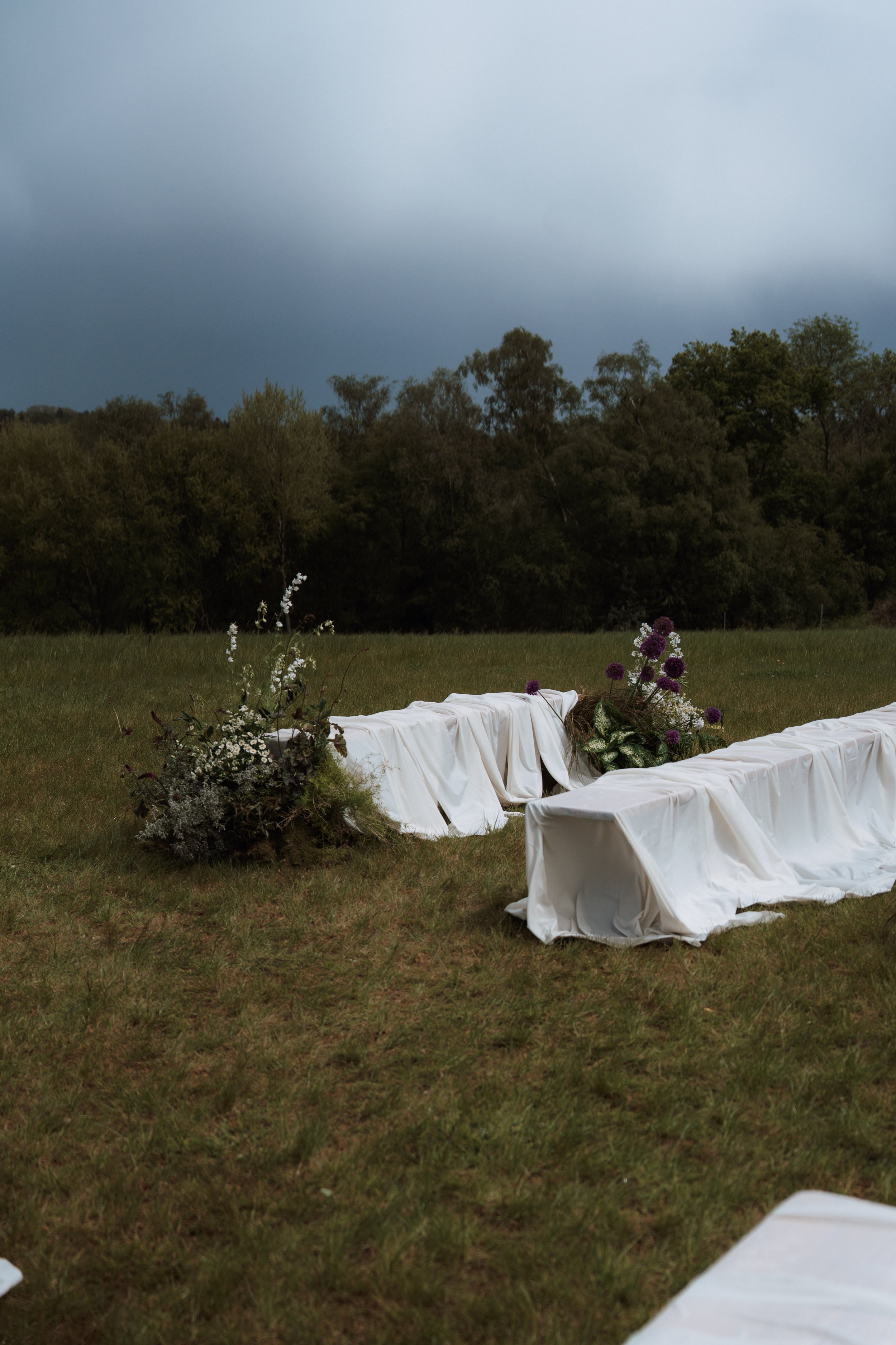 An elegant outdoor wedding reception layout in a grassy field, featuring low tables styled with white fabric draping and wild flowers, set against a backdrop of trees and cloudy skies.