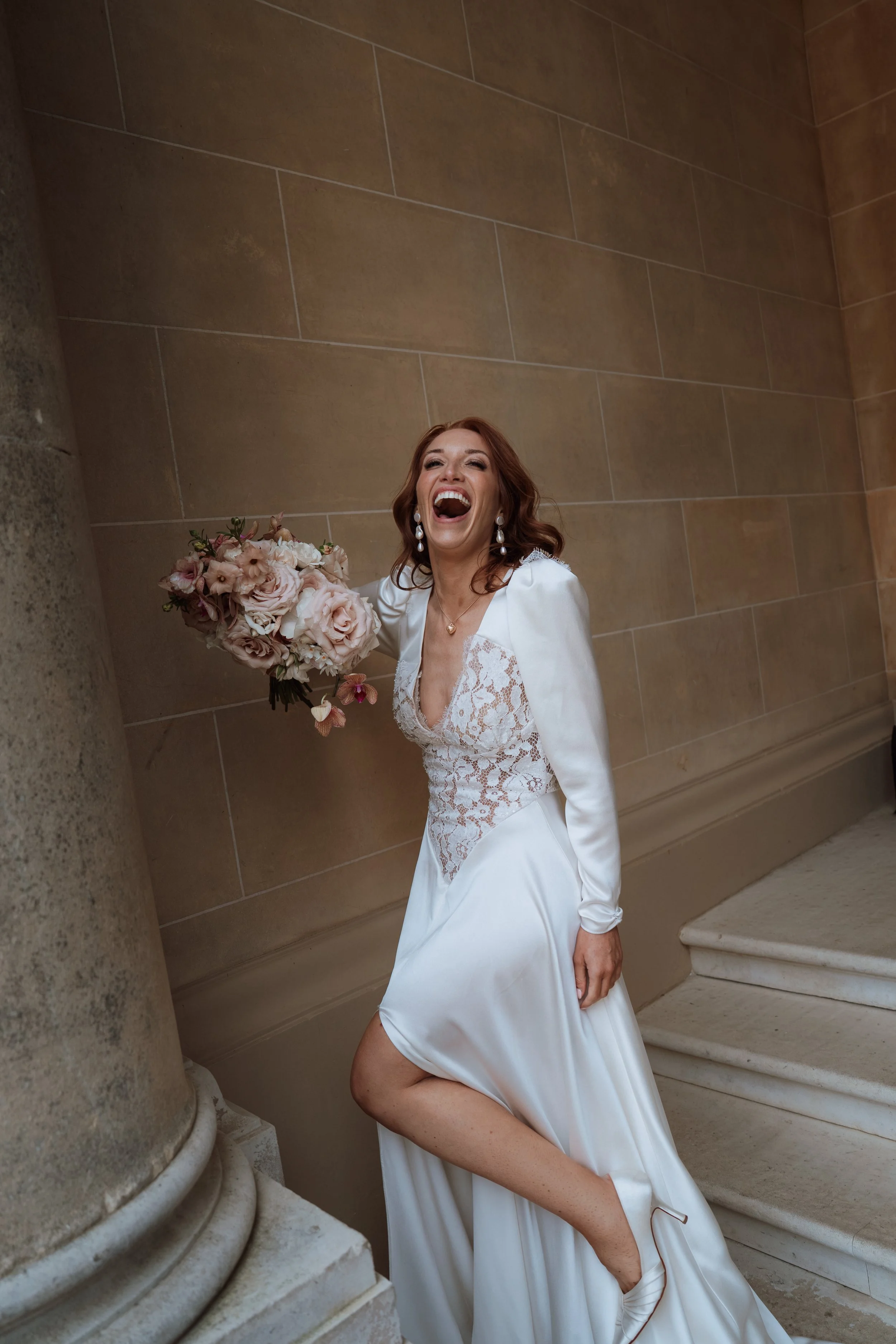 A joyful bride laughing and holding a pink and white bouquet while leaning against a stately stone column next to the grand staircase at Hedsor House.