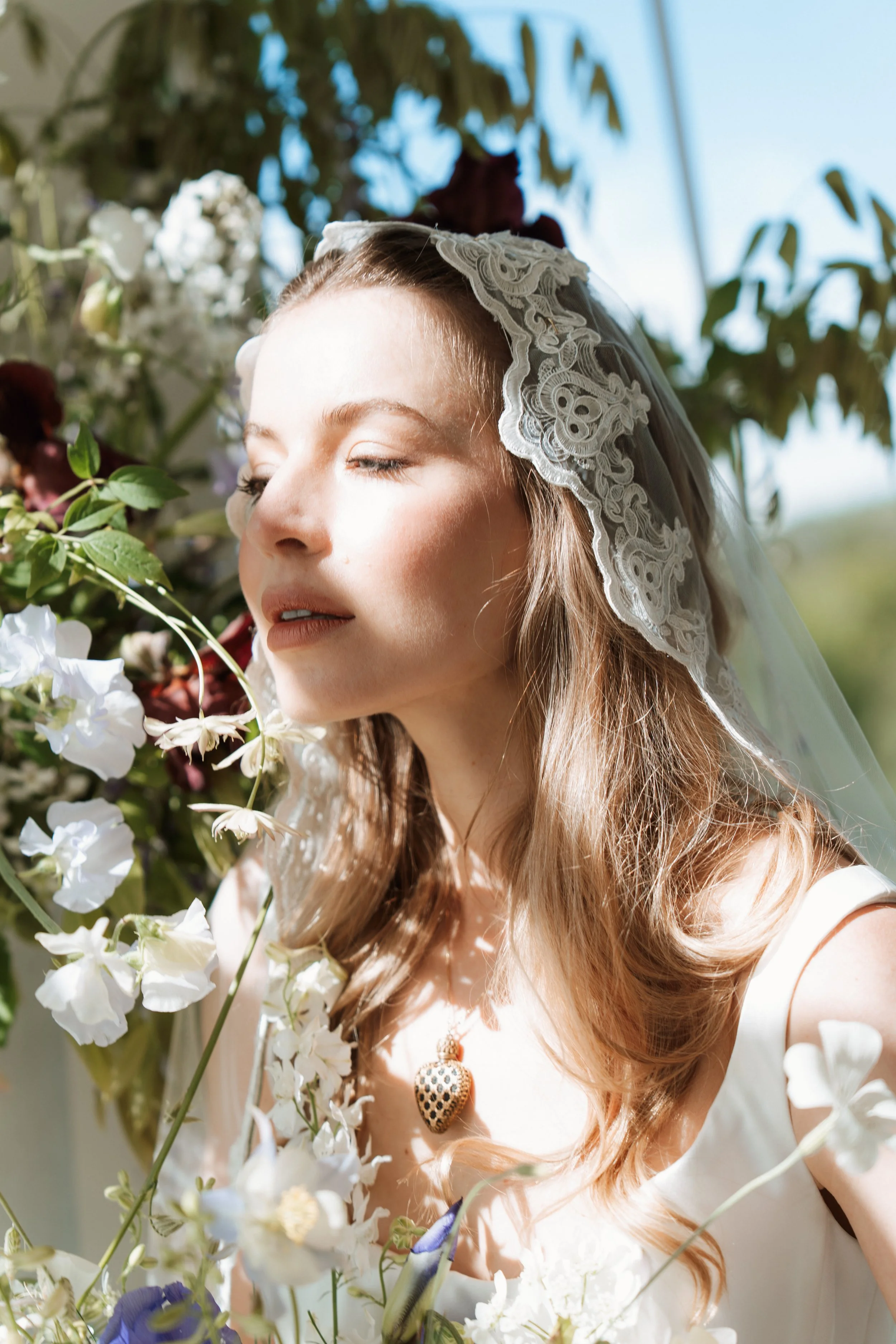 Serene outdoor portrait of a bride with closed eyes wearing a Savannah Miller Bridal wedding dress and delicate lace veil, surrounded by lush greenery and purple flowers.