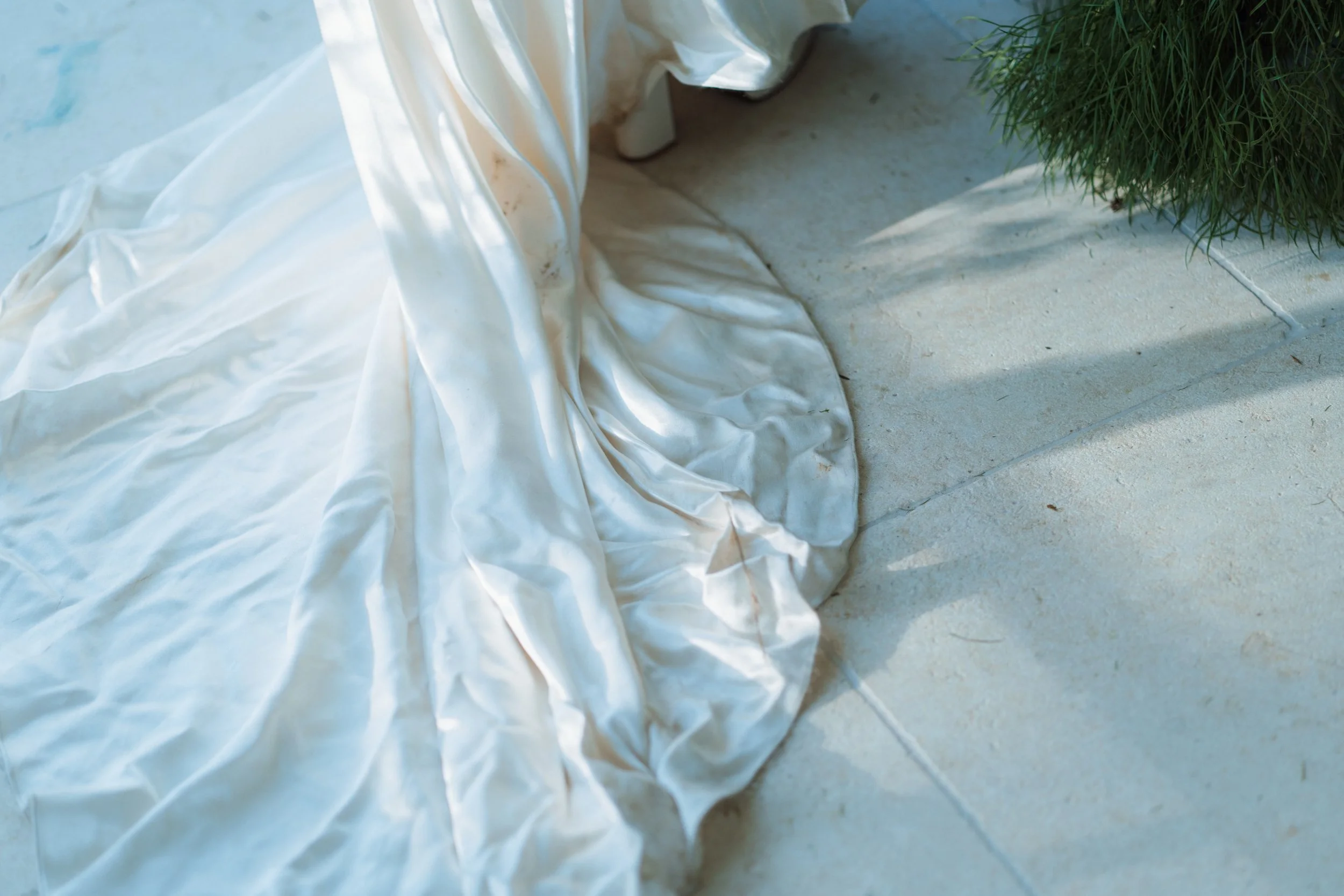 Luxury wedding dress detail shot featuring the elegant white satin train of a Danielle Frankel bridal gown dramatically spread across a concrete floor outdoors next to green foliage.