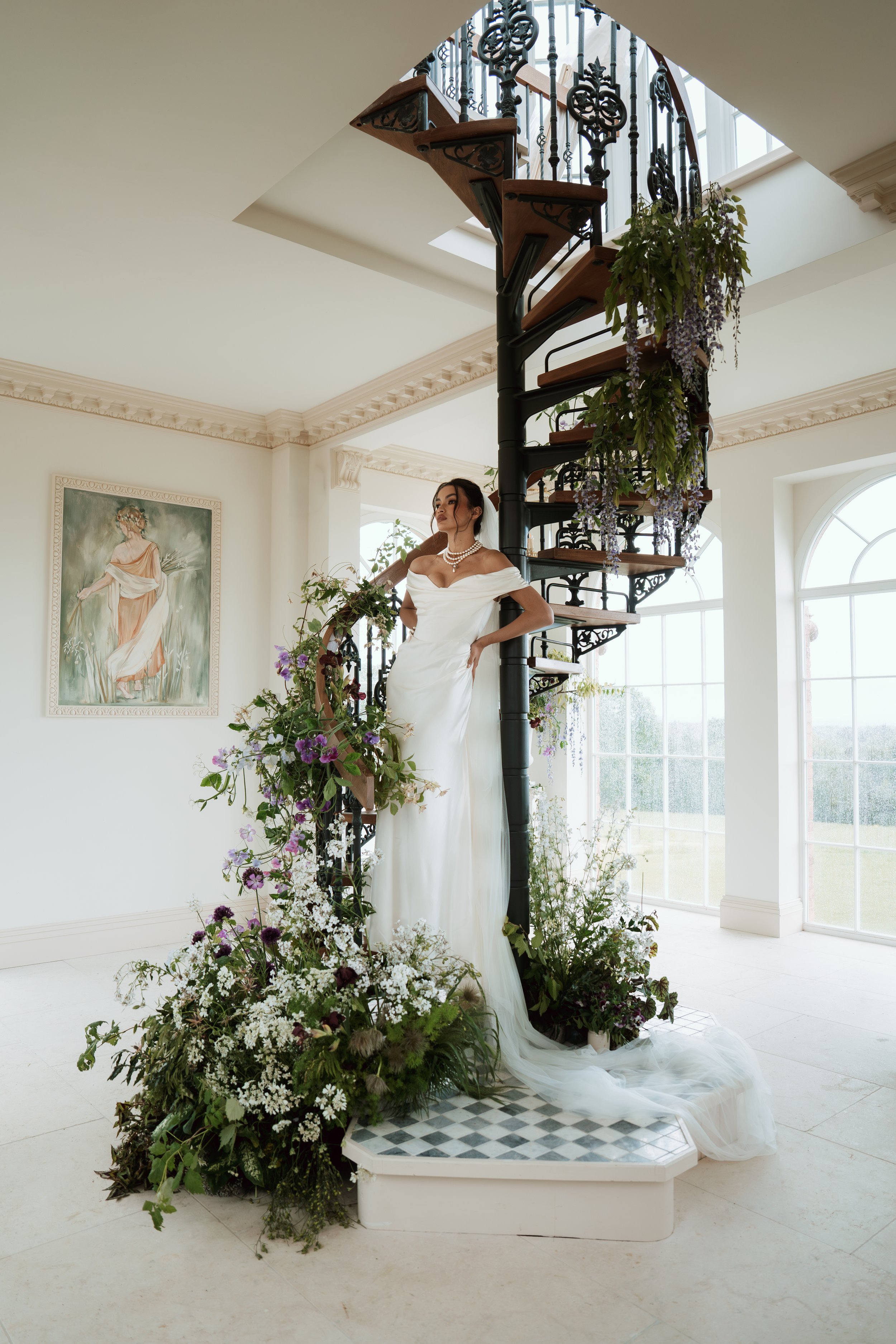 An elegant bride in an off-the-shoulder Vivienne Westwood wedding dress and pearl necklaces stands on a spiral staircase adorned with lush purple and white flowers inside The Folly at Greentrees Estate.