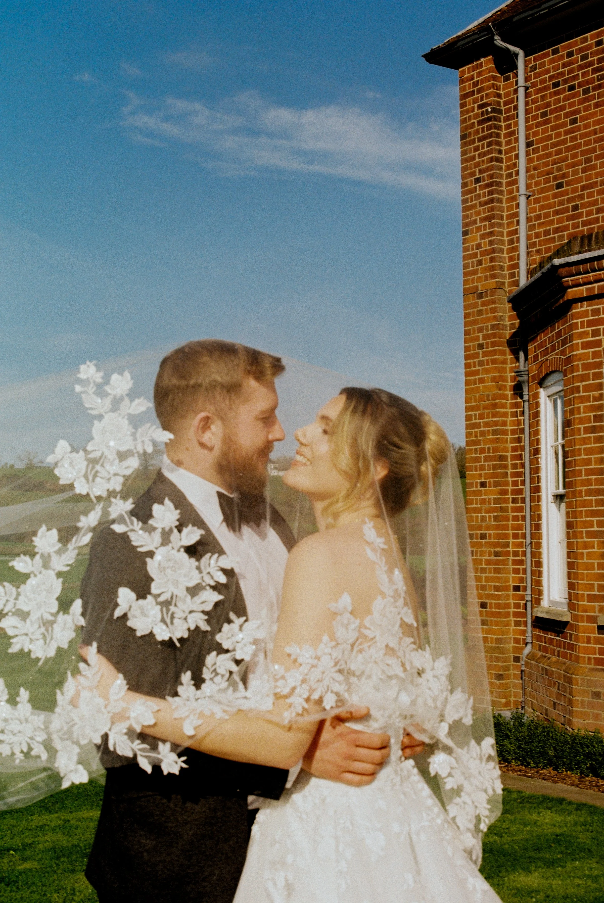 A happy couple in wedding attire, holding each other and smiling, outdoors in front of Downham Hall.