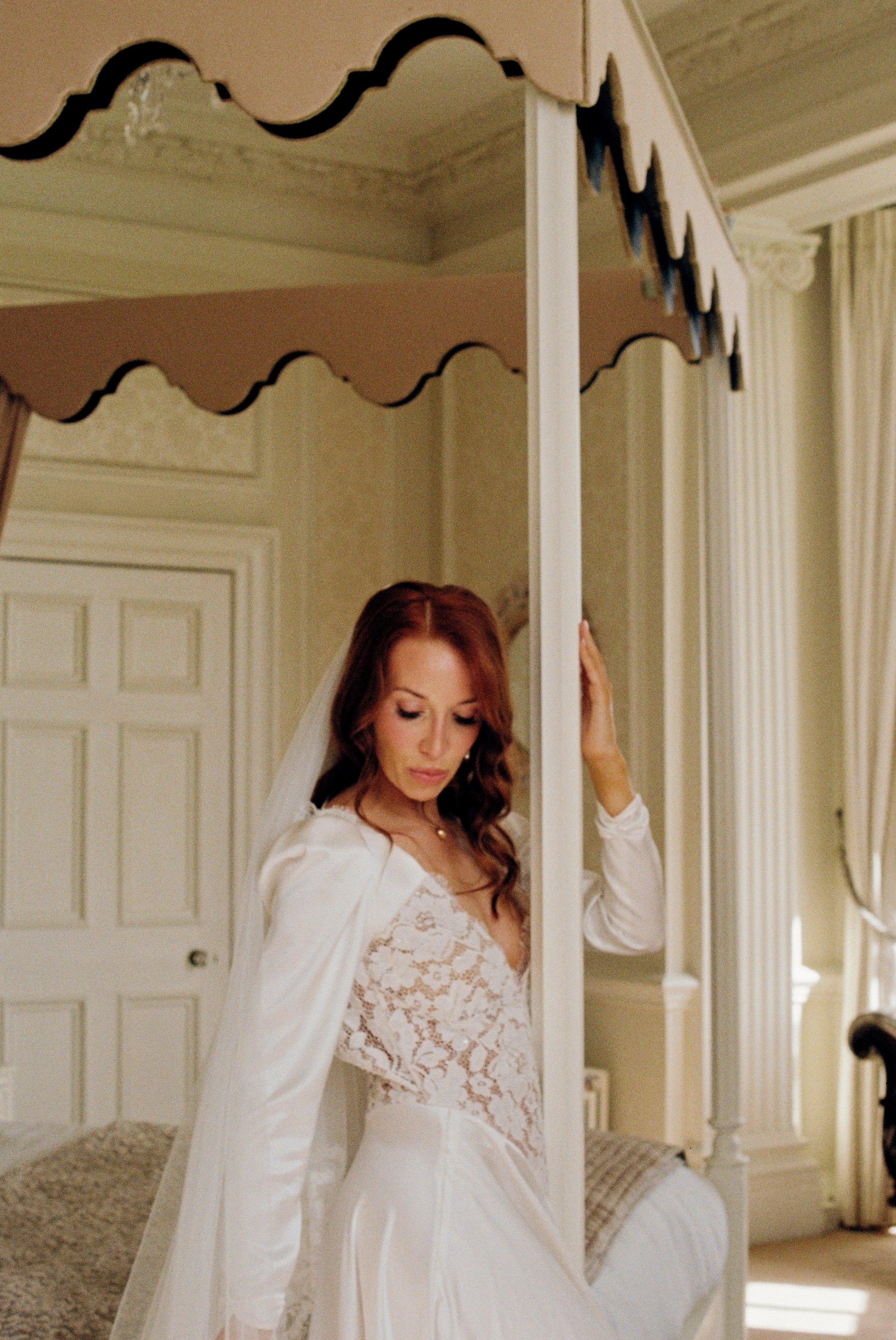 A bride with long red hair in a wedding dress and veil is brushing her hair in front of a mirror.
