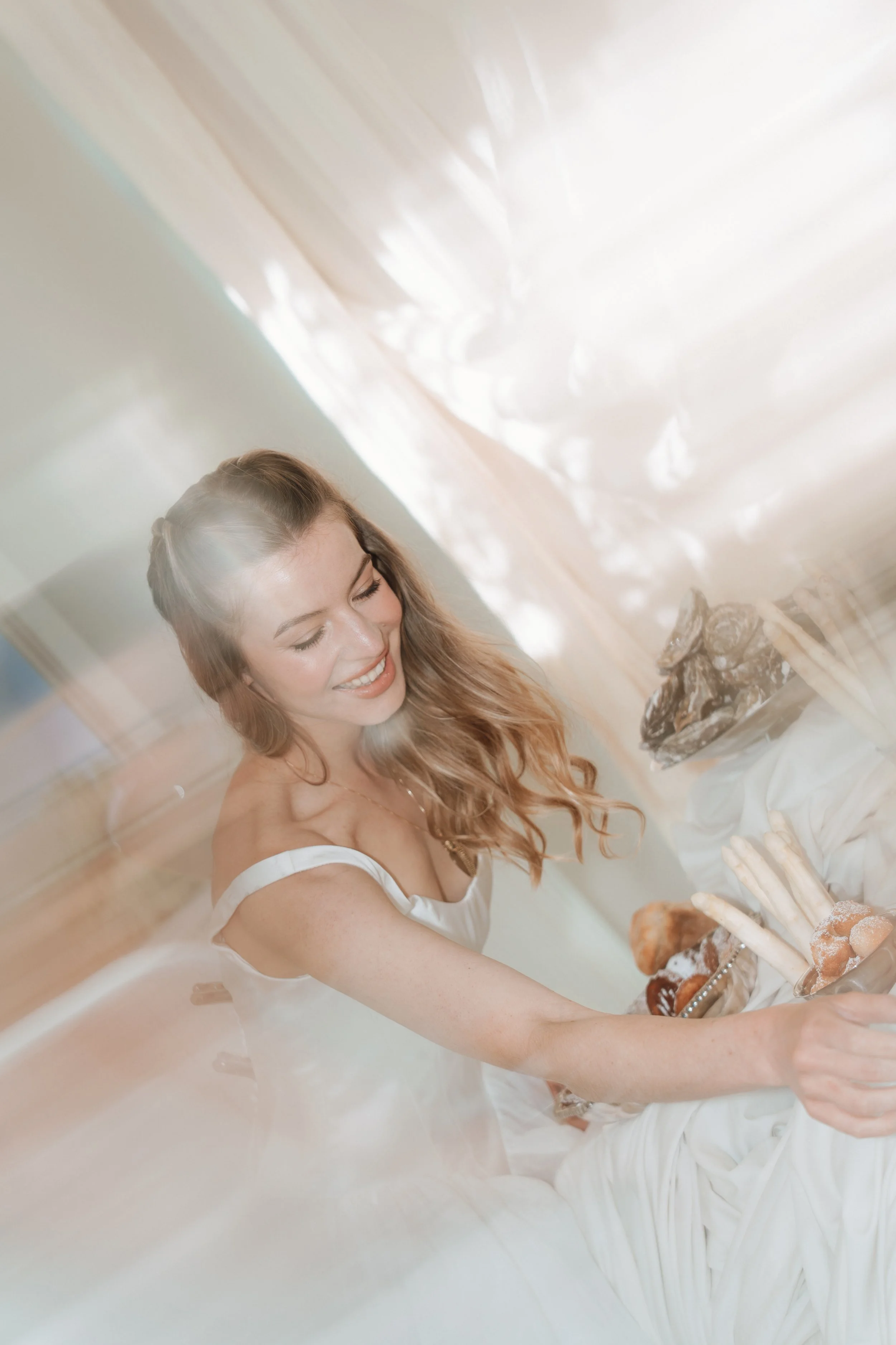 A smiling bride with long, wavy hair wearing a Savannah Miller Bridal wedding dress, reaching for luxury food on a beautifully styled reception table.