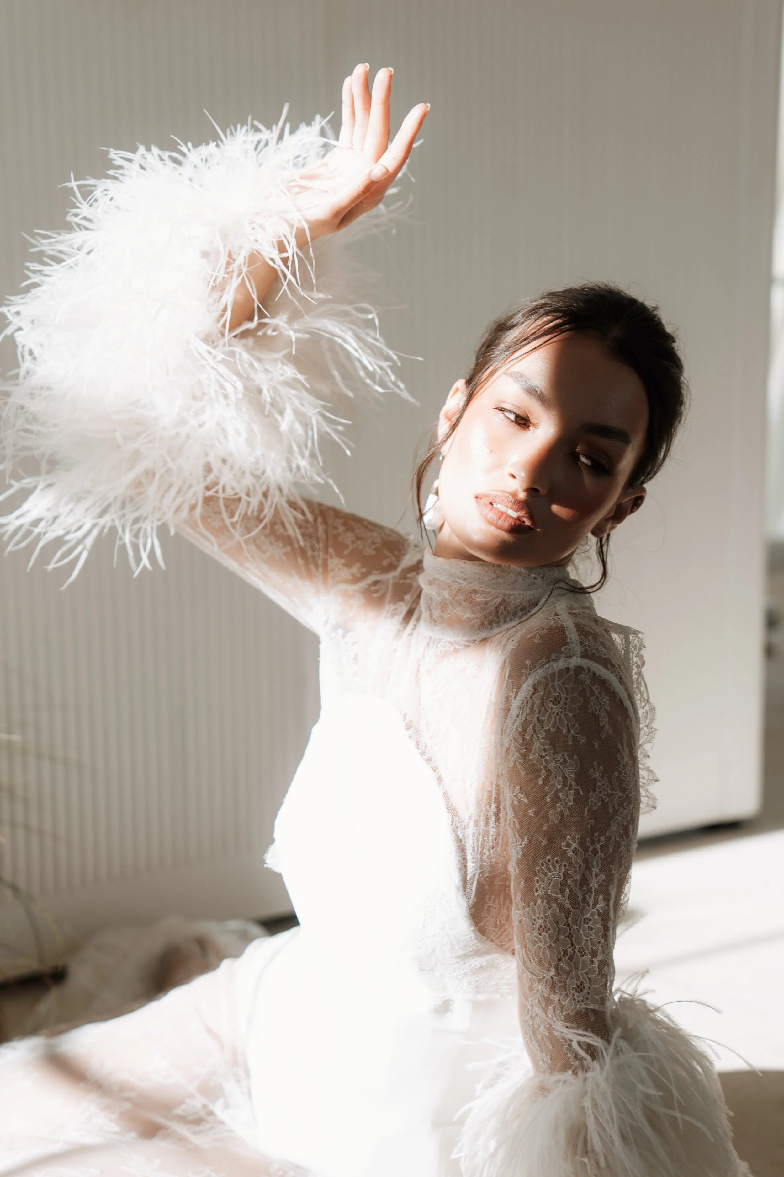 Editorial wedding portrait of a bride in a white lace Asar London dress with statement feathered cuffs, posing gracefully on a bed with natural sunlight and dramatic shadows.