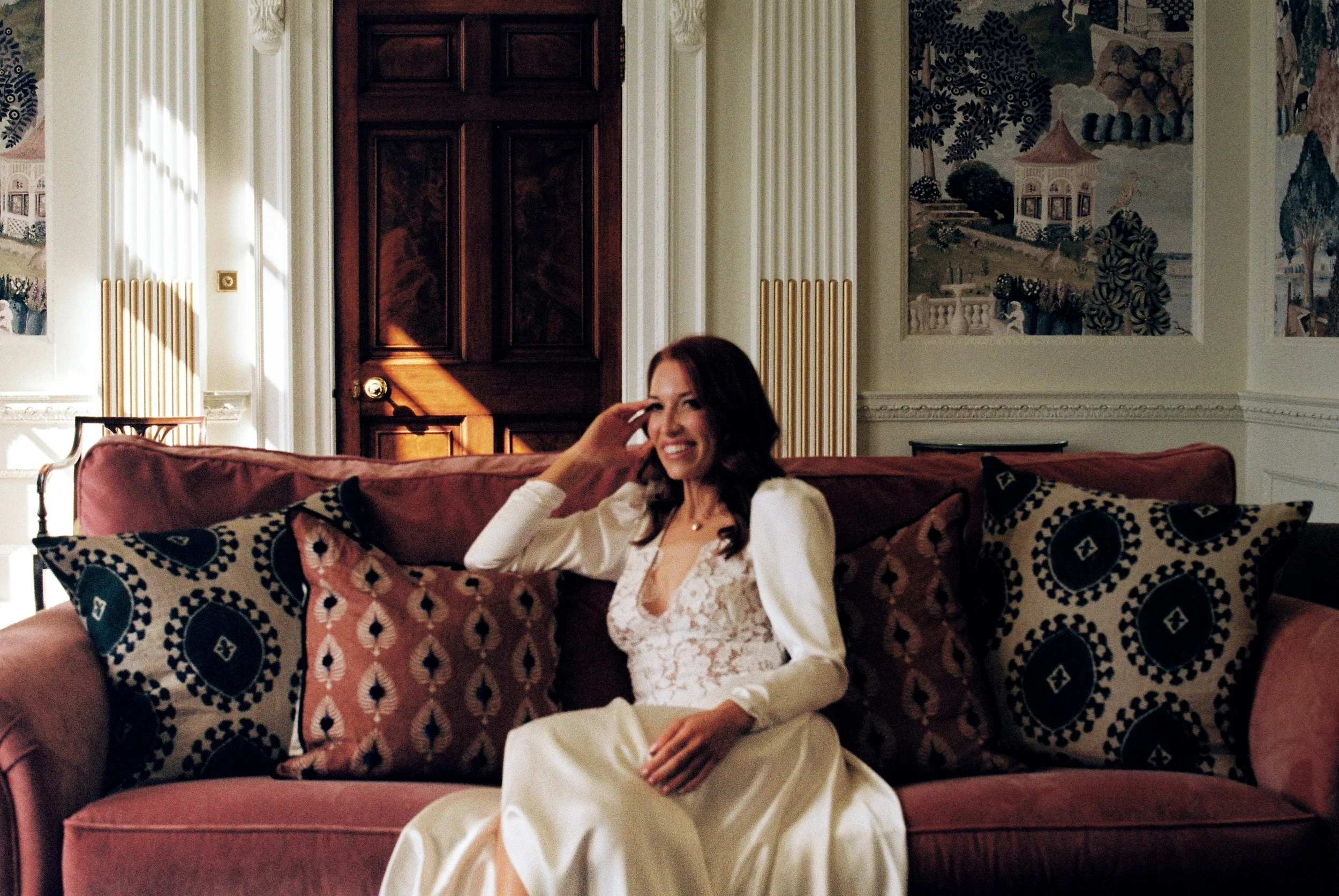 A bride with dark hair smiling and sitting on a pink sofa in an ornate room with large paintings on the wall, a wooden door, and decorative columns.