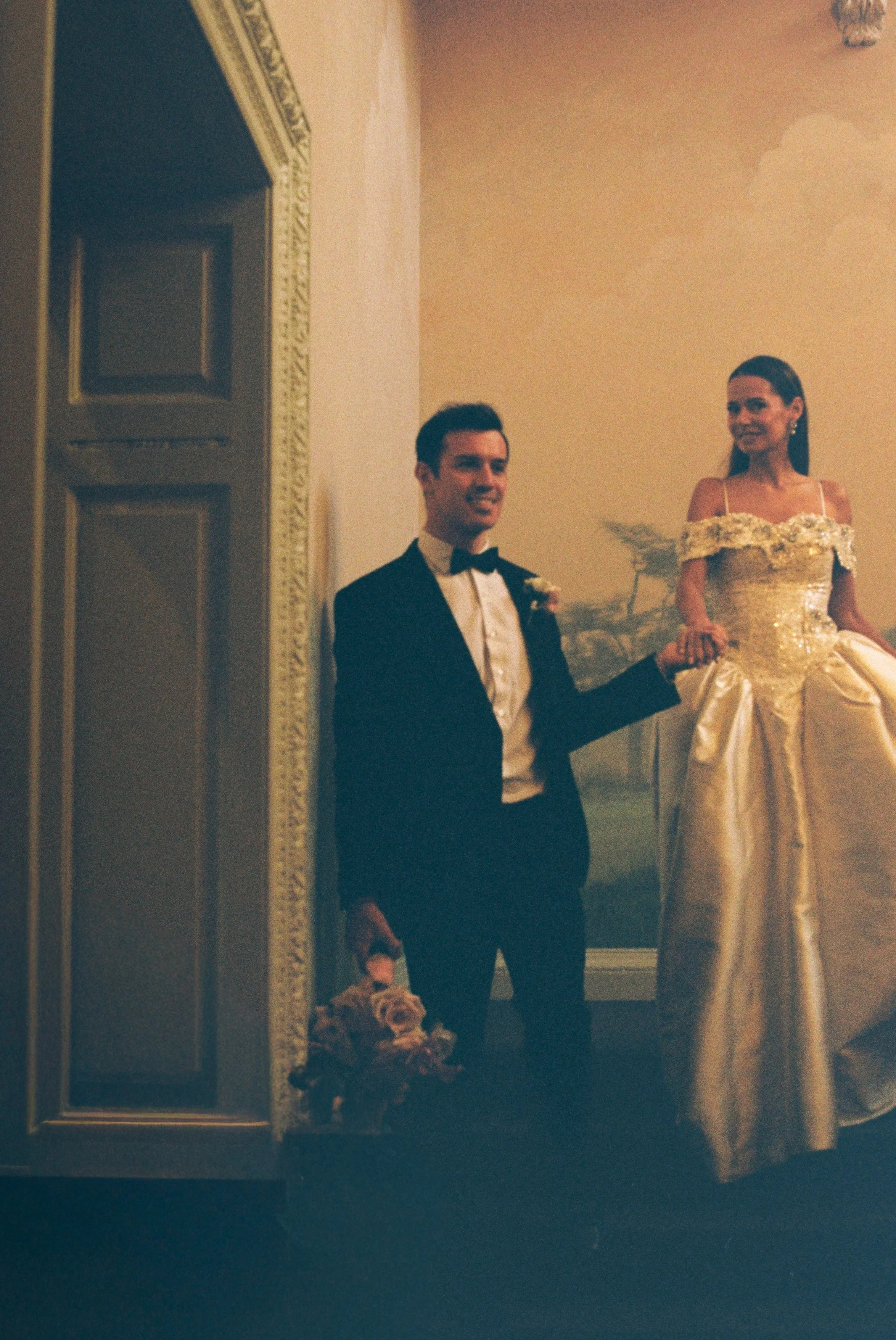Bride and groom at Hedsor House on the main staircase, the man is wearing a tuxedo with a bow tie, and the woman is wearing an ornate gown. They are holding hands.