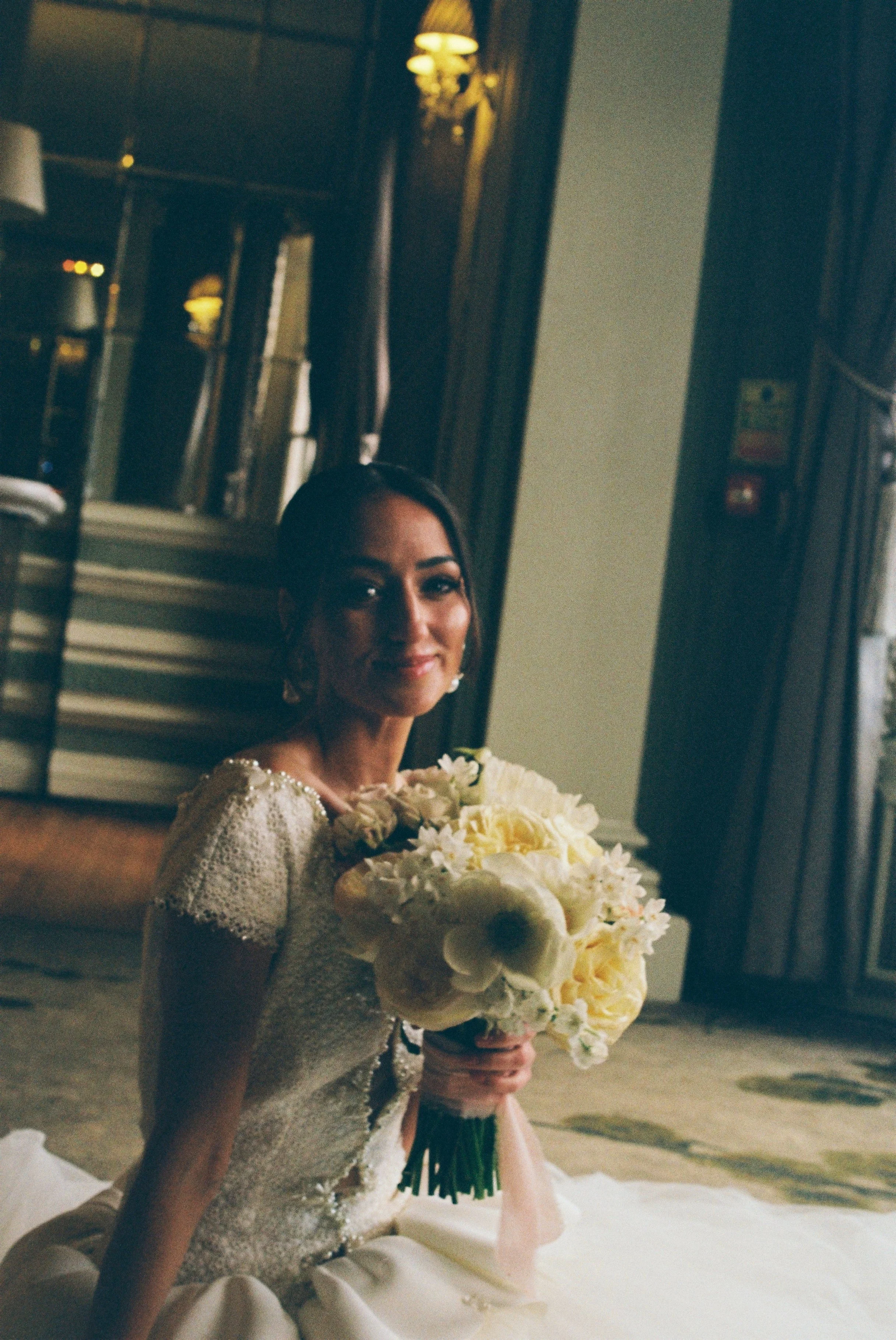 A bride in a bridal dress holding a bouquet of white and cream flowers, sitting indoors near a mirror and a window, with warm lighting and dark wooden decor.