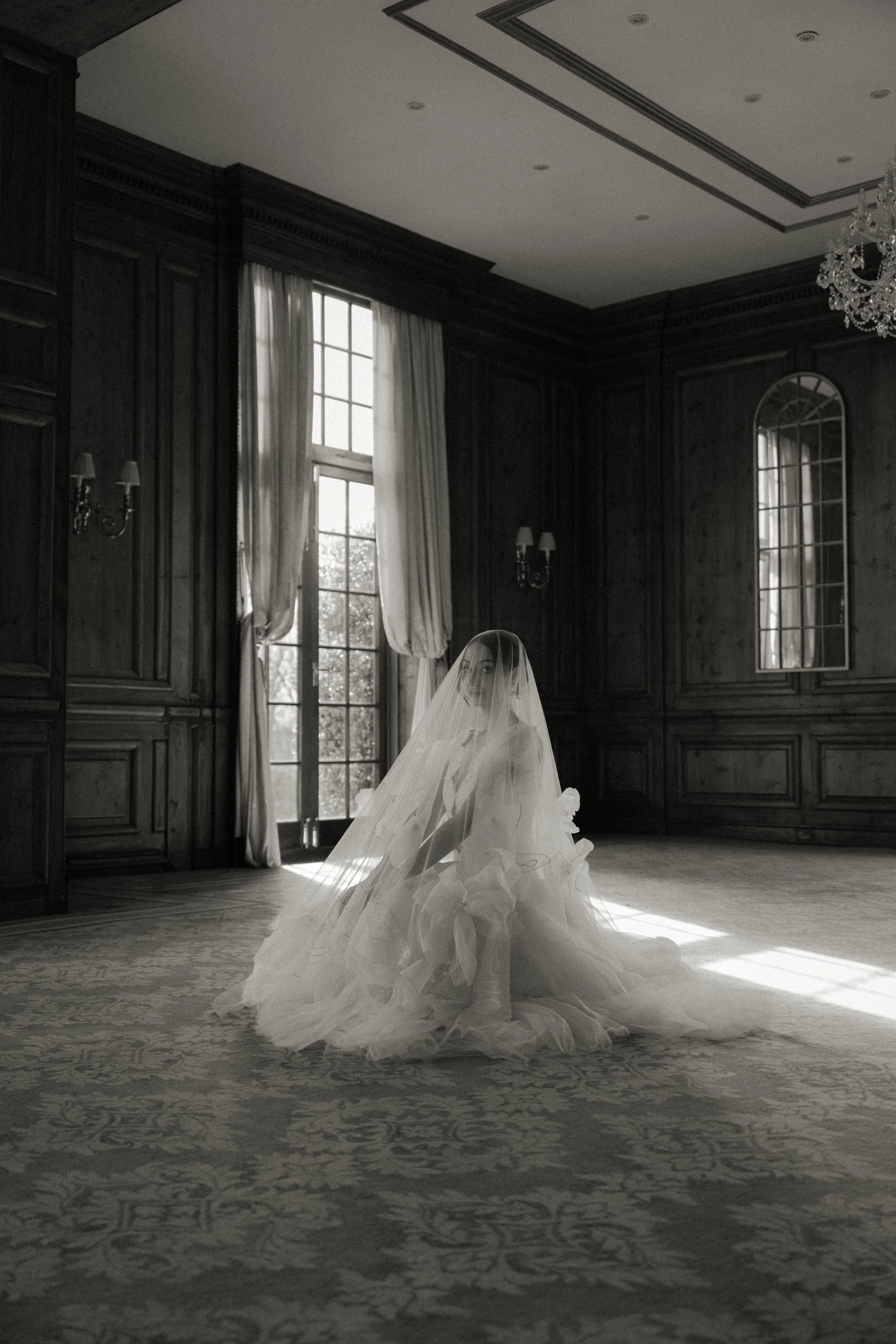 A black and white editorial bridal portrait of a bride kneeling gracefully under a full wedding veil on an ornate carpet inside the grand ballroom at Hedsor House, captured by Buckinghamshire wedding photographer Jacs Batchelor.