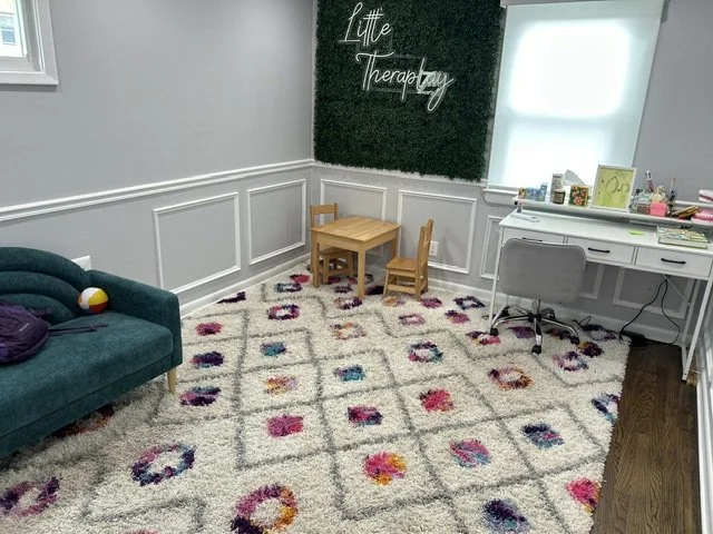 A therapy room with a green couch, a small wooden table with two chairs, a white desk with a chair, and a wall with a neon sign that says 'Little Therapy.' The room has a colorful patterned carpet and a window with blinds.