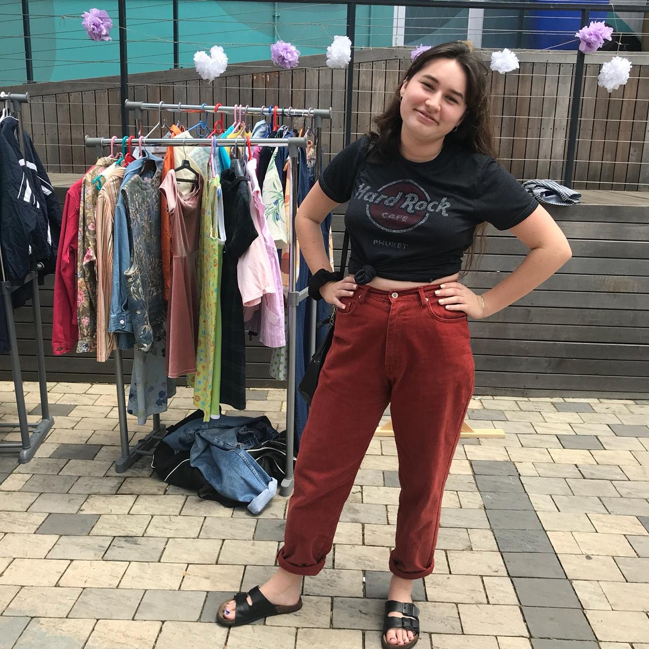 A young woman with dark brown hair wearing a black Hard Rock Cafe Phuket t-shirt, red pants, and black sandals standing smiling with hands on hips next to a clothing rack with various colorful garments, outdoors on a paved area with purple and white paper pom-pom decorations hanging above.