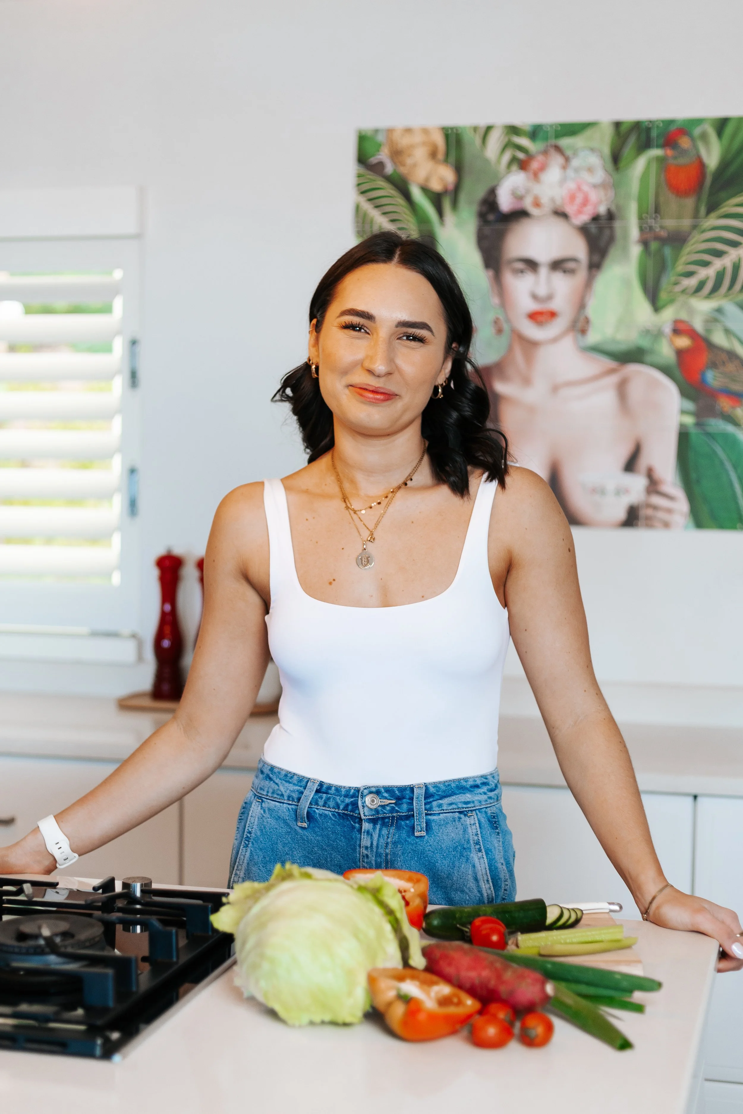 A woman with dark hair, wearing a white tank top and jeans, stands in a kitchen in front of a countertop with fresh vegetables, including lettuce, cucumber, and cherry tomatoes. Behind her, a colorful poster of Frida Kahlo with tropical leaves and parrots is hanging on the wall.