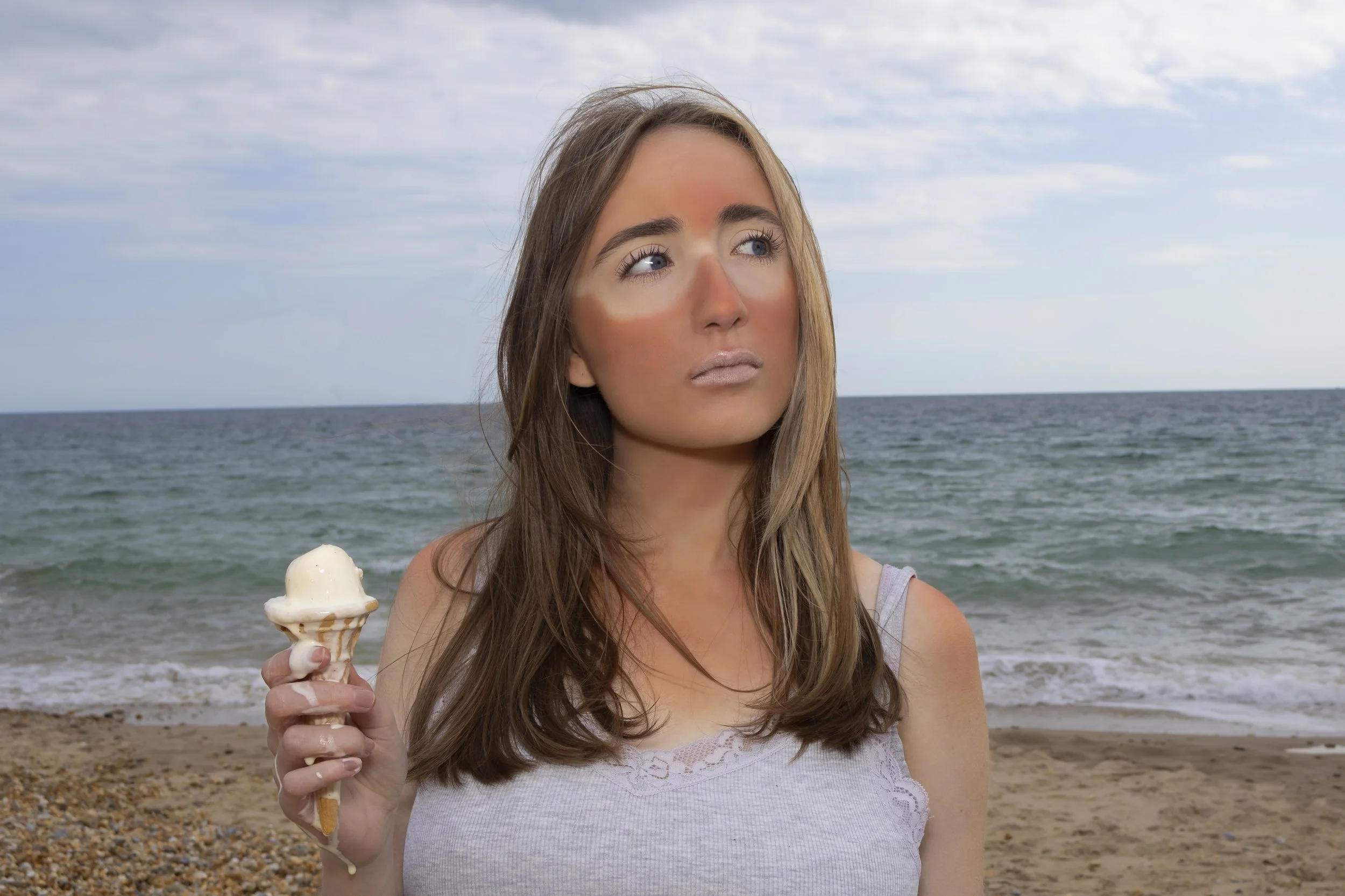 A woman with long brown hair holding a vanilla ice cream cone on a beach, with the ocean and sky in the background.