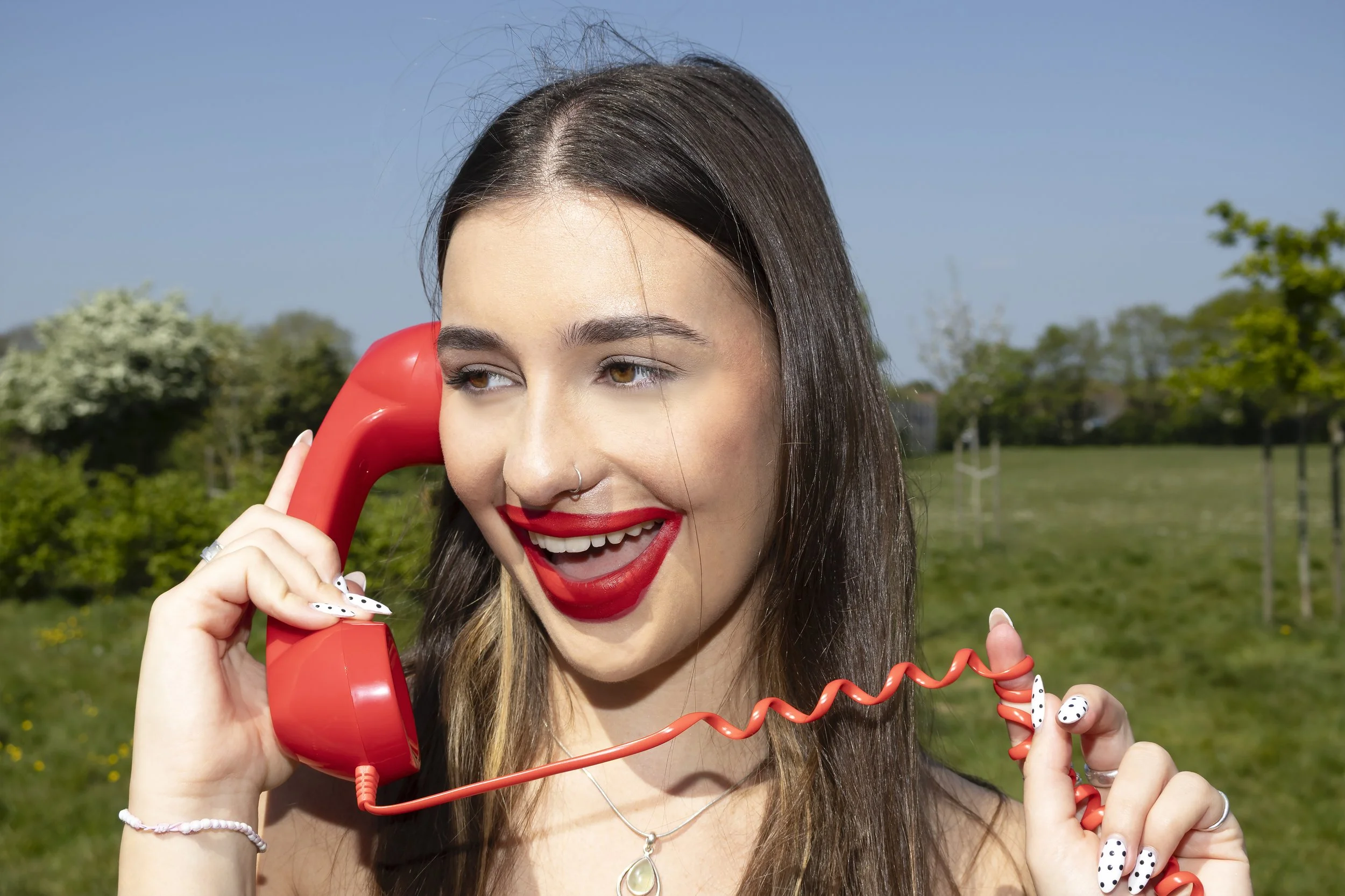 Young woman with dark brown hair, red lipstick, and a nose ring, holding a red telephone receiver to her ear outdoors with a grassy field and trees in the background.