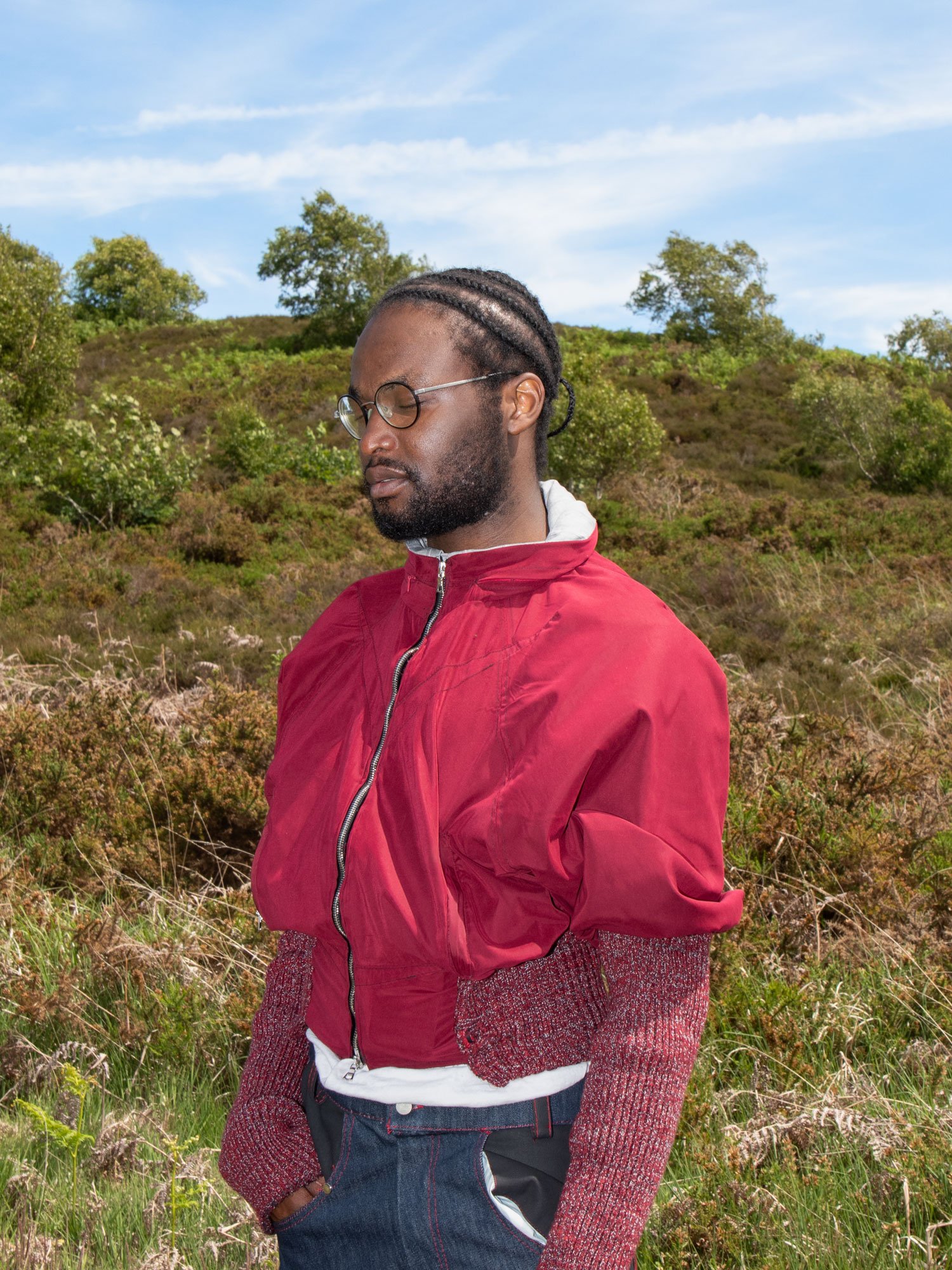 A man with glasses and braided hair, wearing a red jacket over a white shirt, standing outdoors in a natural landscape with hills and trees under a blue sky.