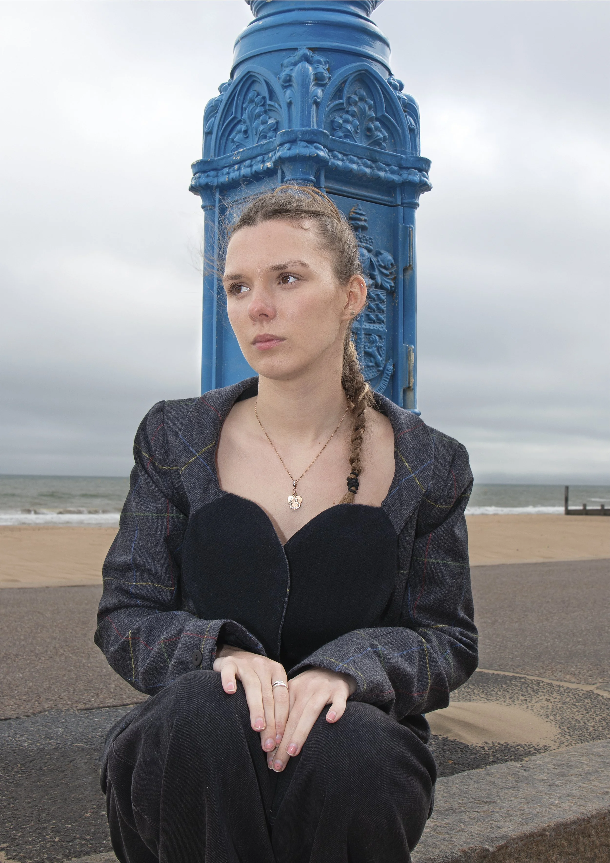 A woman sitting cross-legged outdoors near the beach with her hands clasped on her knees, wearing a dark coat and black pants, with a blue sky and ocean in the background.