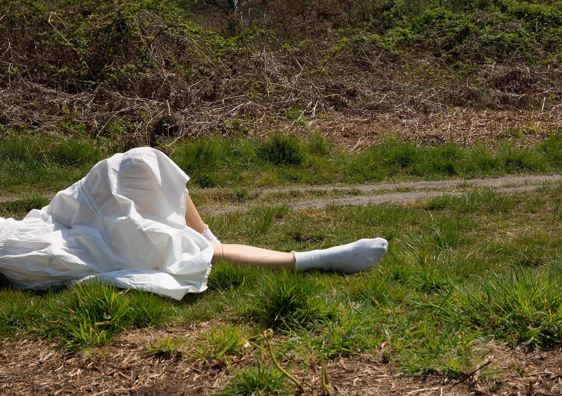 Person lying on the grass in front of a hedge, wearing a white dress and white socks.