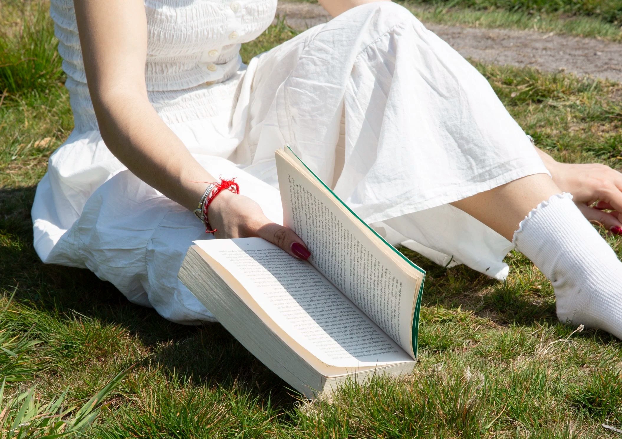 Person sitting on grass reading a large open book outdoors with green grass and a dirt path in the background.