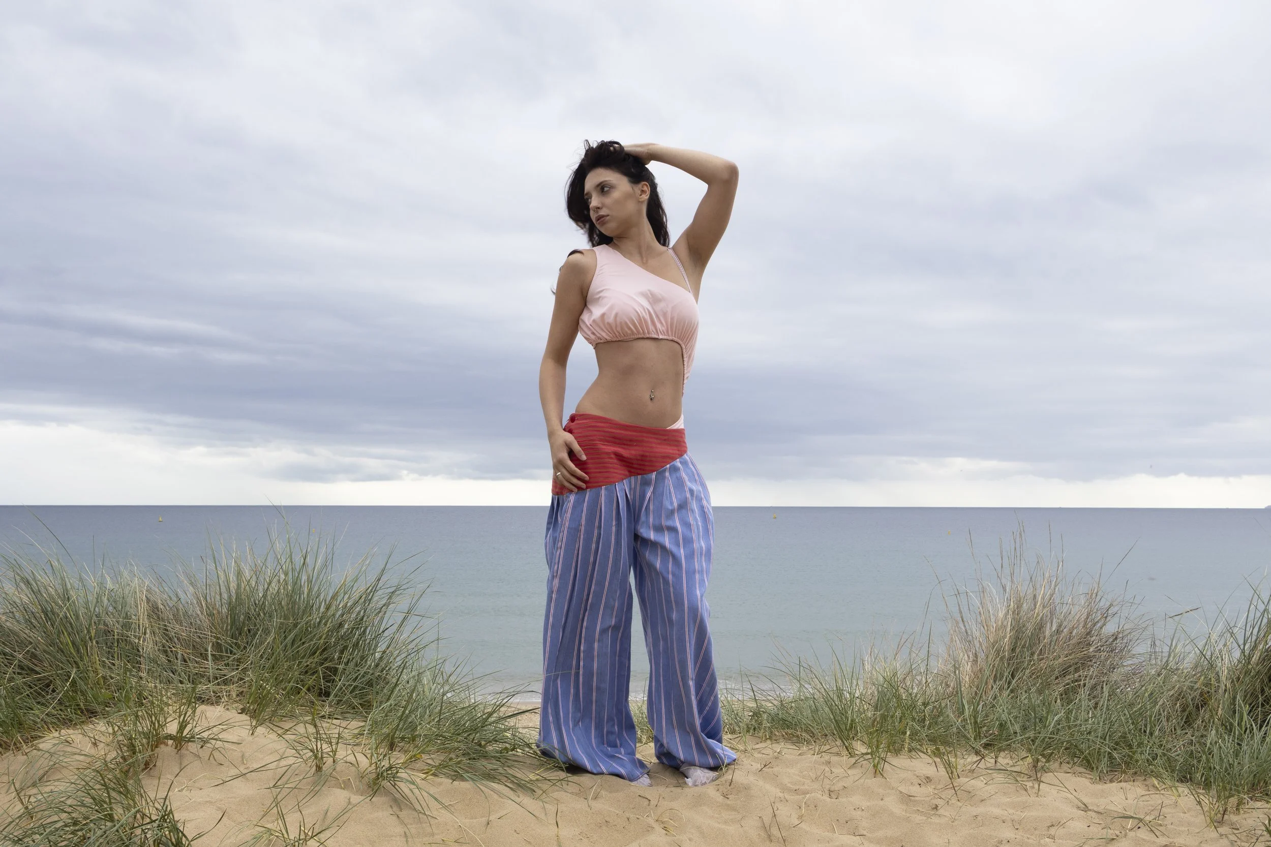 Woman standing on sandy beach with tall grass, ocean, and cloudy sky in the background, wearing a pink top and loose striped pants.