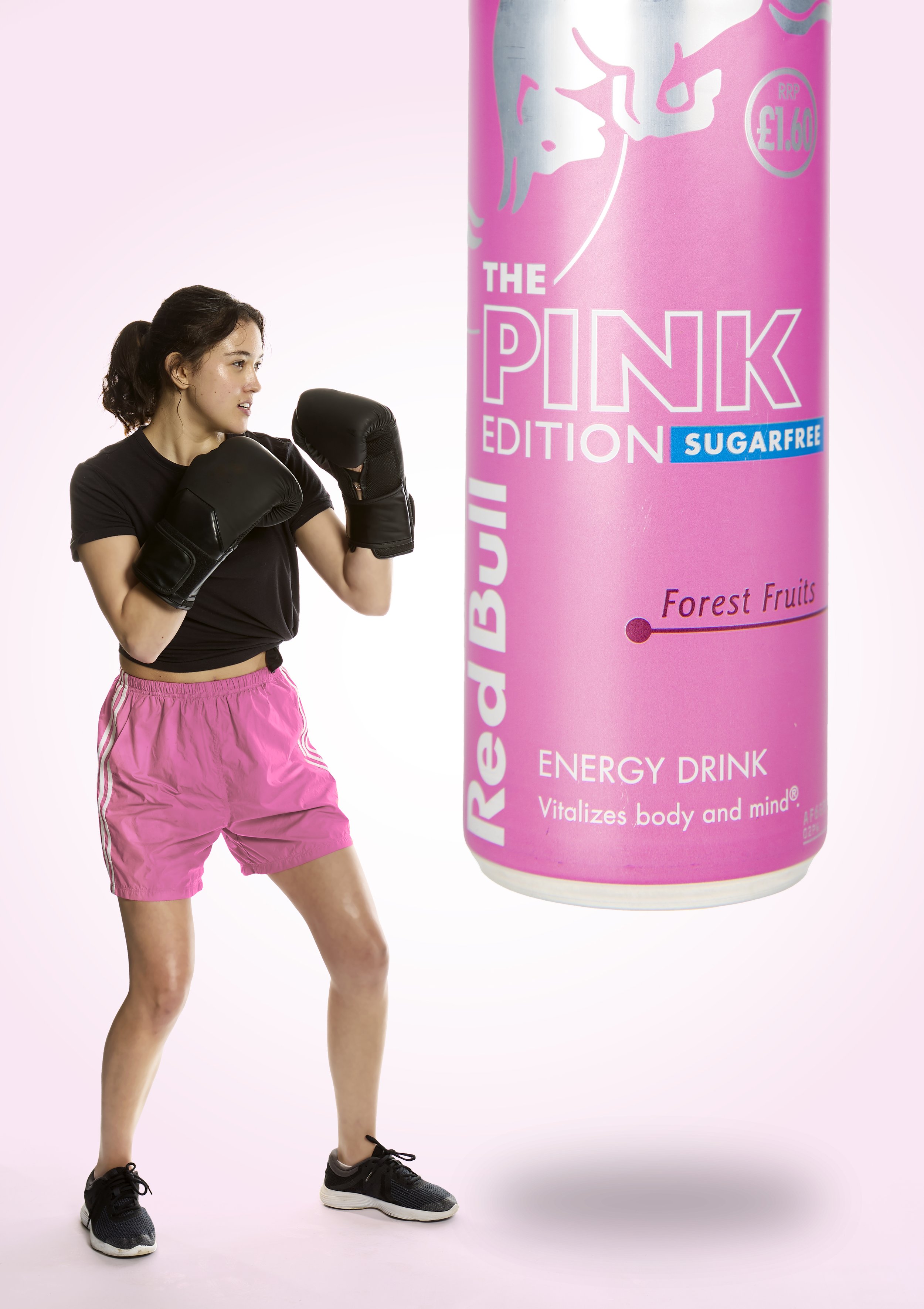 A young woman in pink shorts, black t-shirt, and black athletic shoes boxing with pink boxing gloves in front of a large can of energy drink labeled 'The Pink Edition Sugar-Free' with a flavor note of 'Forest Fruits' against a light pink background.