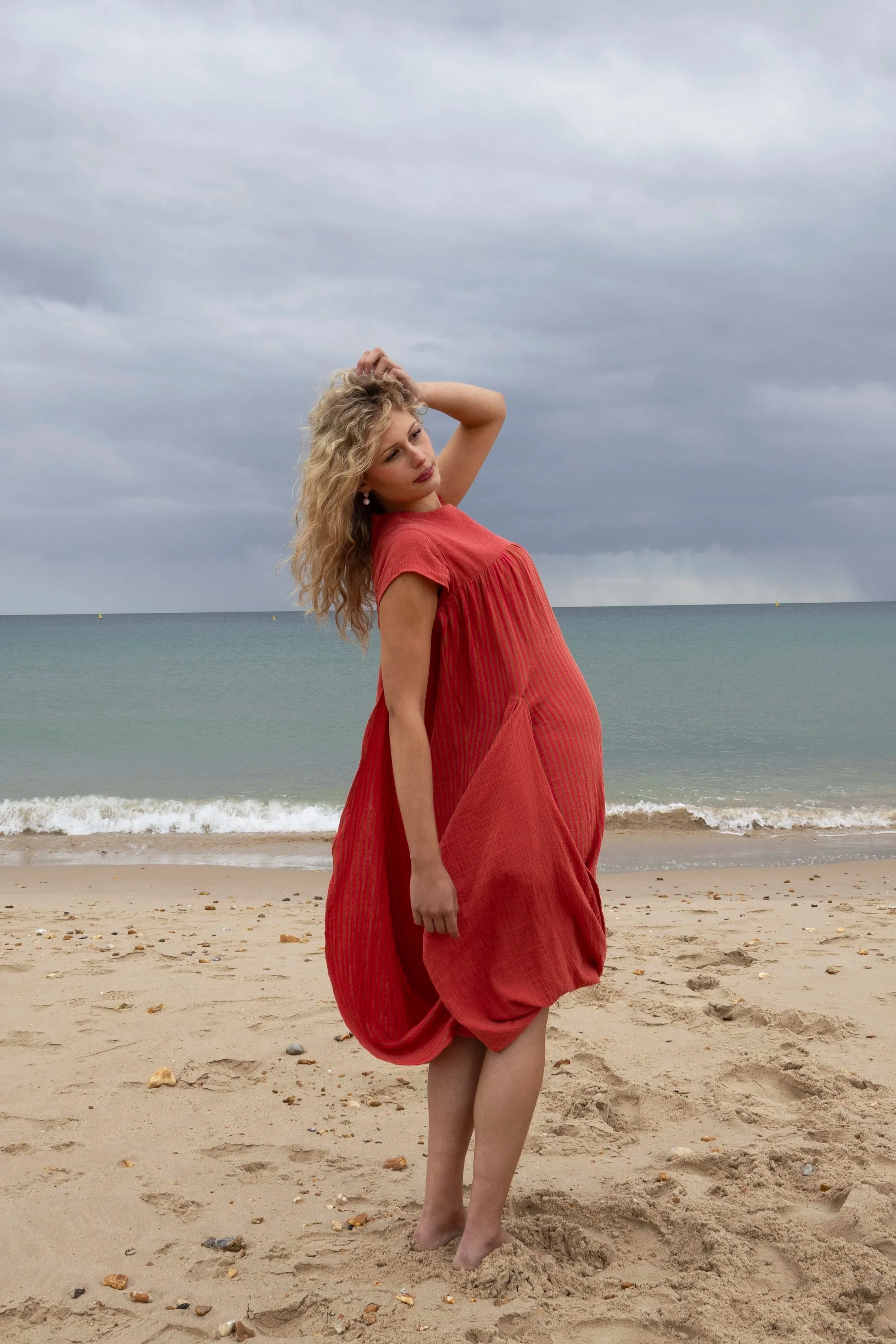 Woman with curly blonde hair in a red dress standing on a sandy beach with rocks, overcast sky, and ocean in the background.