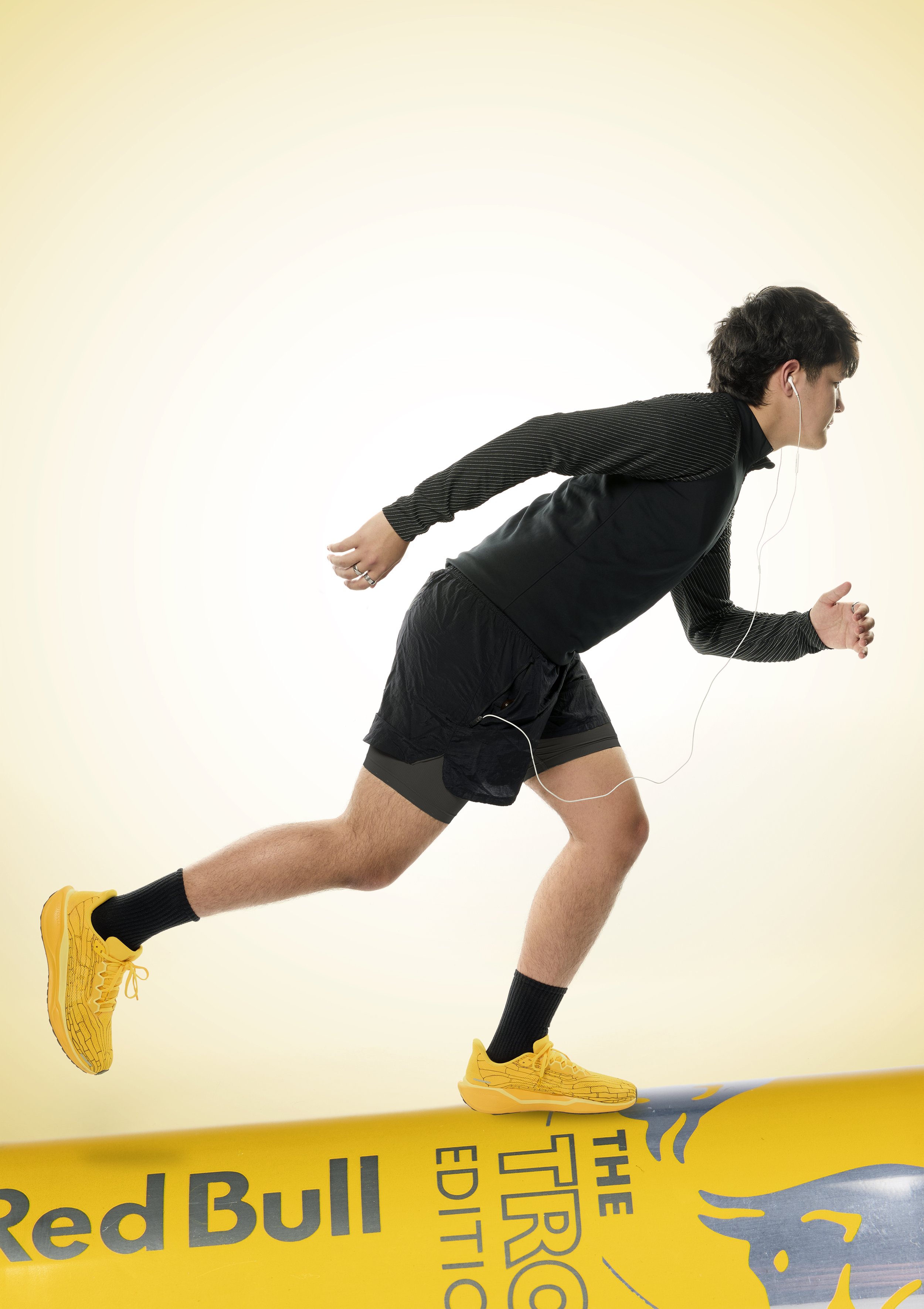 A man dressed in running gear including a black jacket, black shorts, yellow athletic shoes, and black socks, is running on a treadmill. He has earphones in and is in a slight forward-leaning running posture.