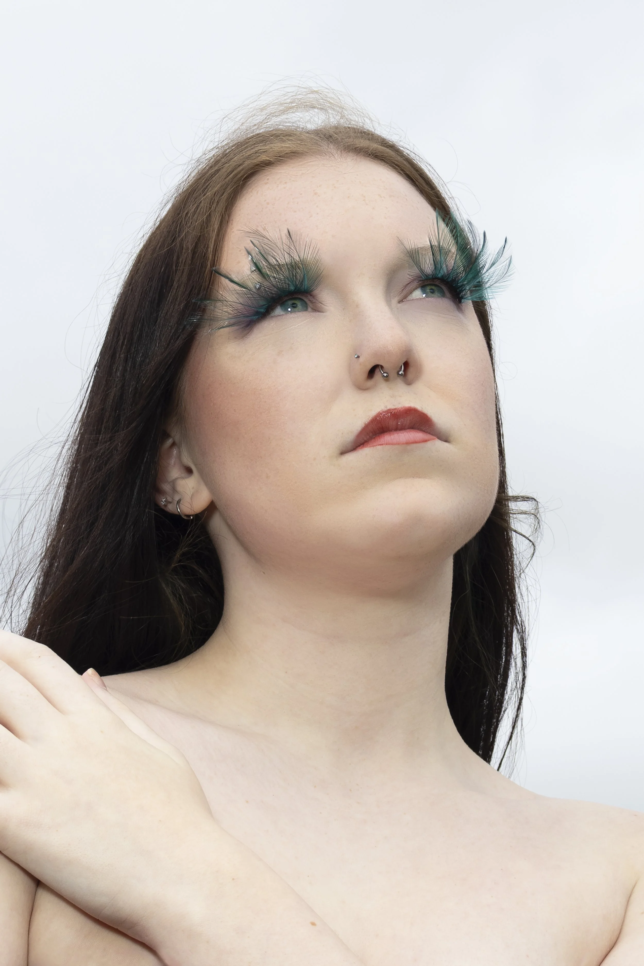 A woman with long dark hair and light skin, wearing dramatic teal feathered false eyelashes, with freckles and red lipstick, appears to be looking upward against a plain light background.