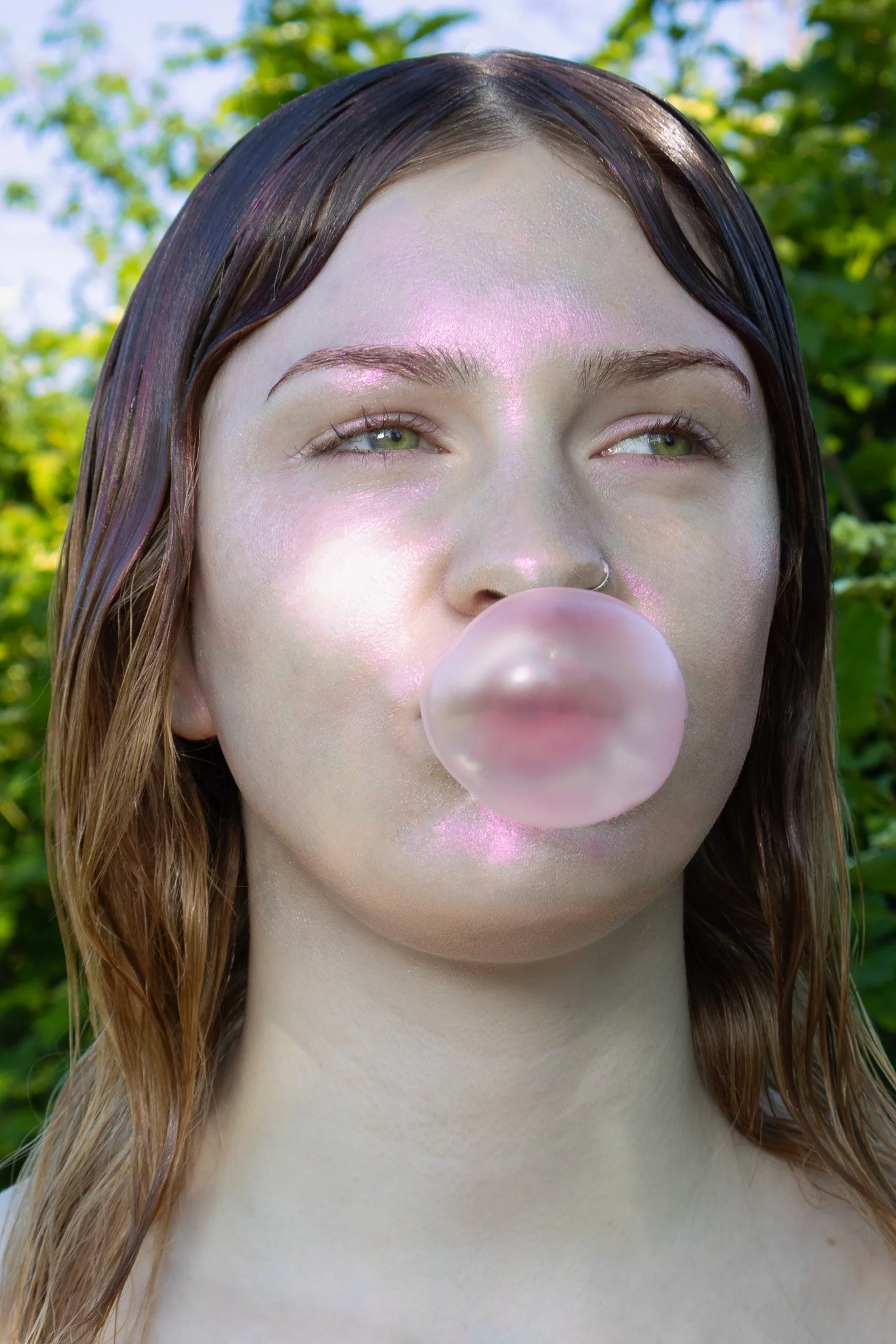 A woman with wet brown hair, light skin, and green eyes is blowing a bubble with pink bubblegum outdoors, with green trees in the background.