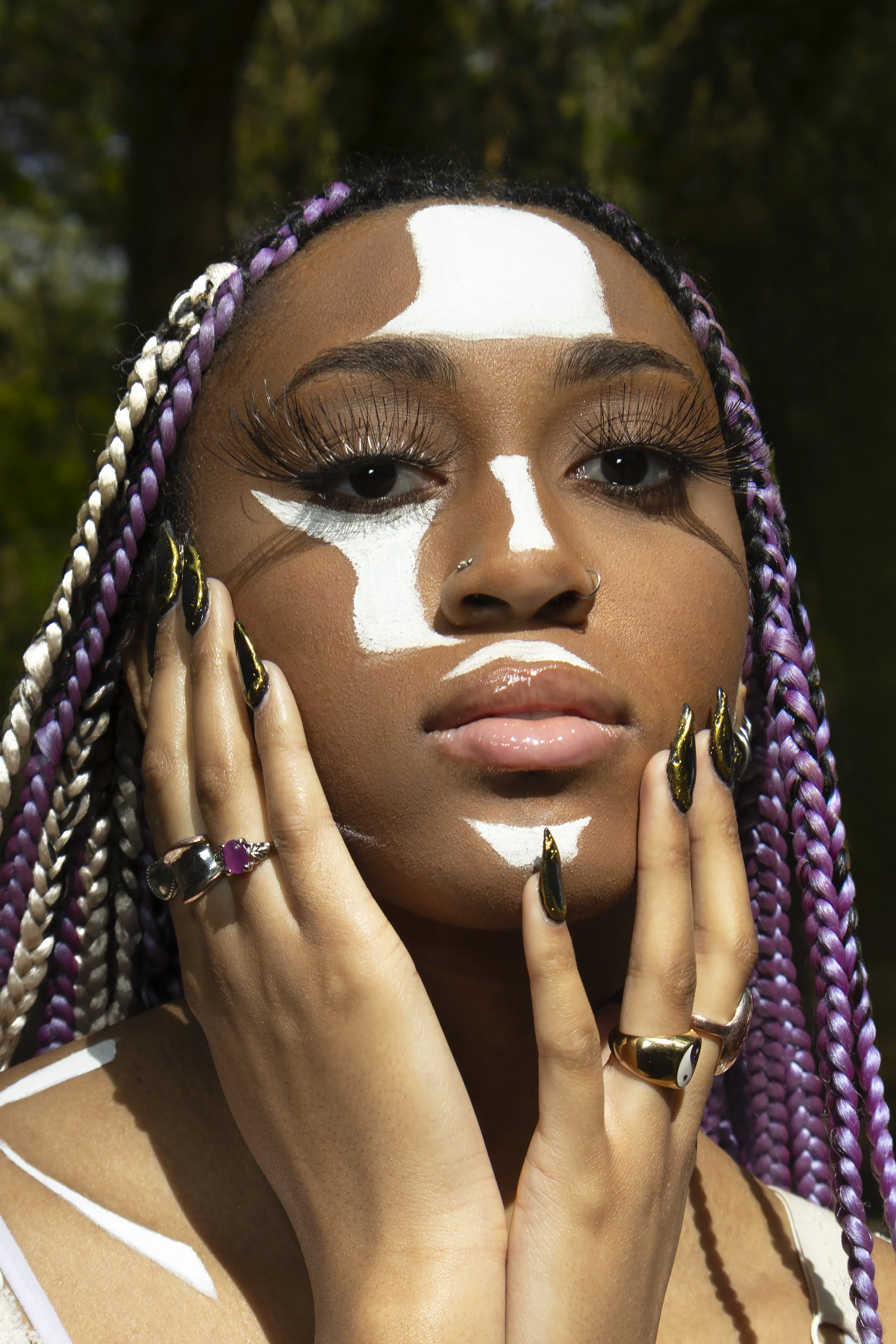 A young woman with purple and white braided hair and gold jewelry touches her face, with white paint on her forehead, nose, and chin, outdoors with trees in the background.