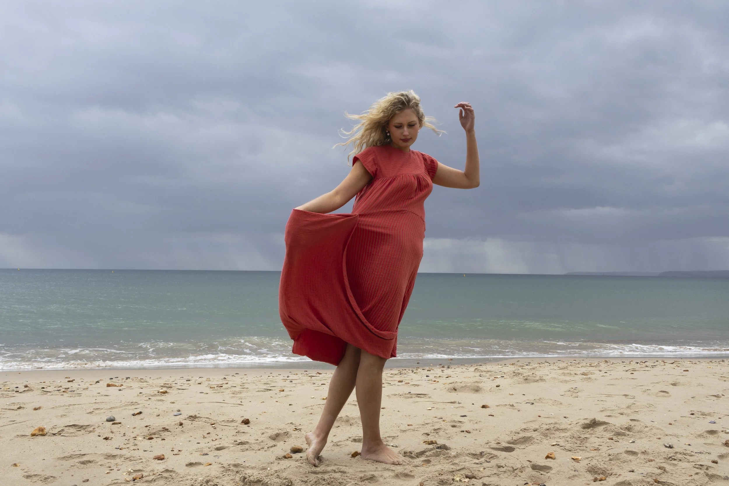 A woman with blonde hair wearing a flowing red dress is standing barefoot on a sandy beach with gentle waves in the background. The sky is overcast with dark clouds.
