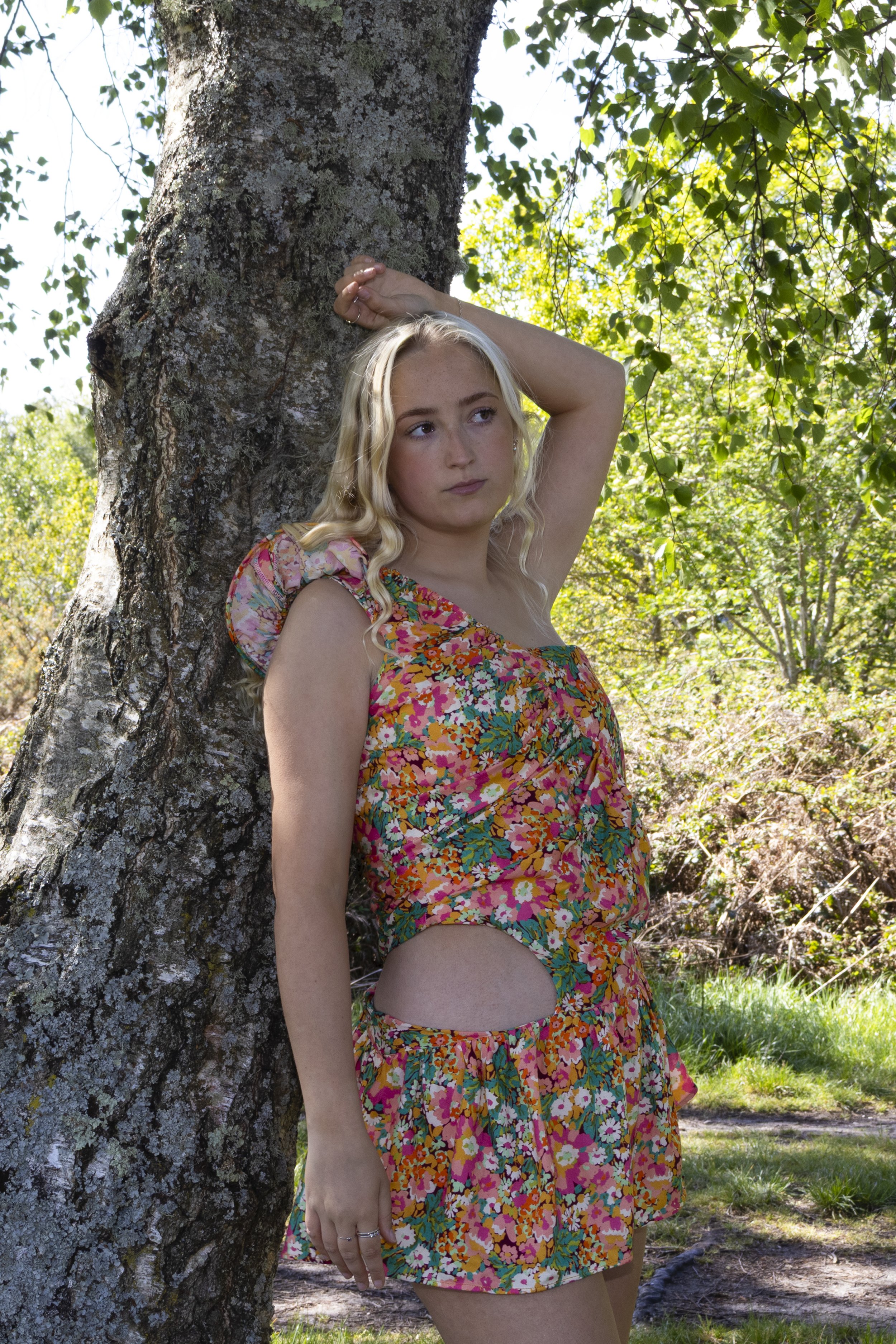 A woman wearing a colorful floral dress with side cutouts poses outside next to a large tree with textured bark, surrounded by lush green leaves and sunlight filtering through.