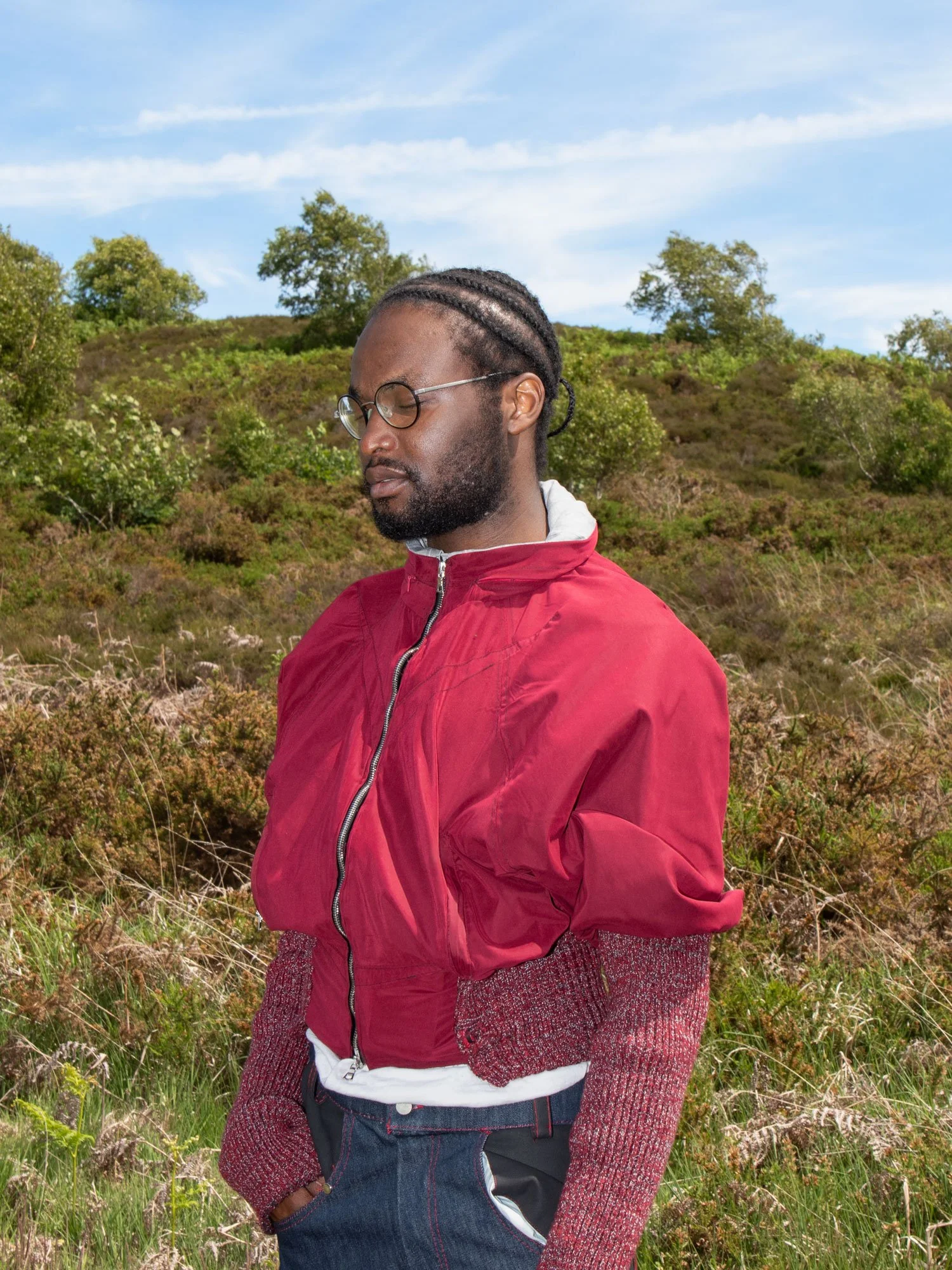 A man with glasses and braided hair standing outdoors in a grassy area with trees and a blue sky in the background, wearing a red jacket and dark pants.