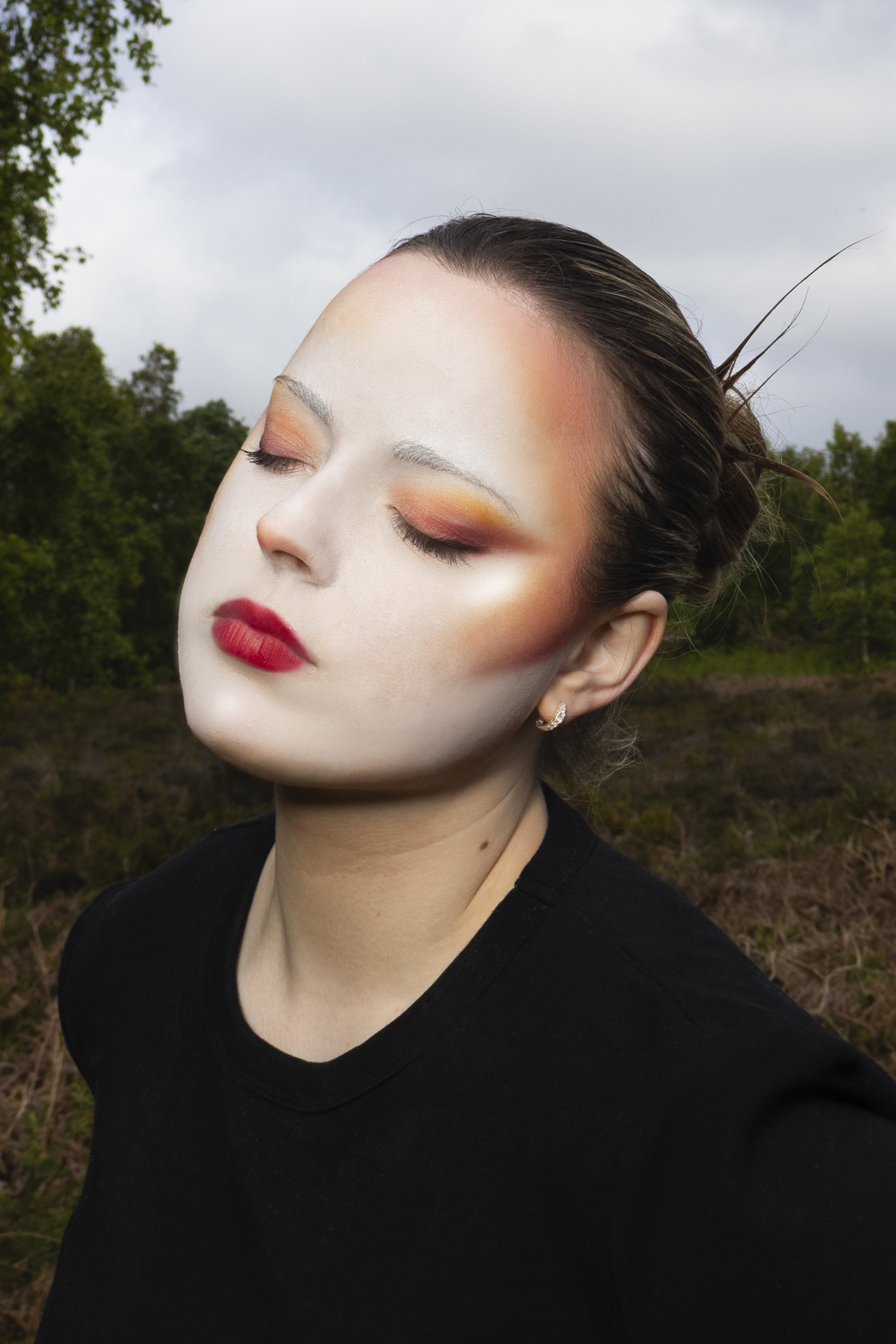 A woman with closed eyes and makeup, wearing a black top, outdoors with trees and a cloudy sky in the background.