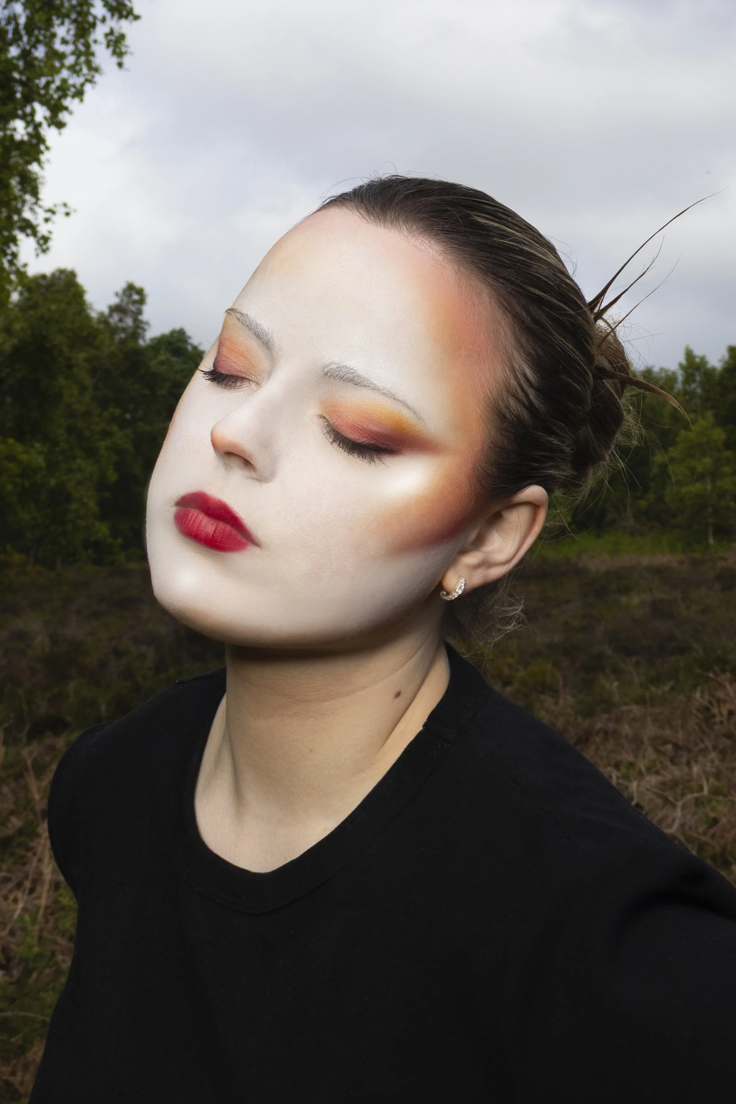Close-up of a woman with makeup and earrings, with her eyes closed, outdoors against a cloudy sky and trees.