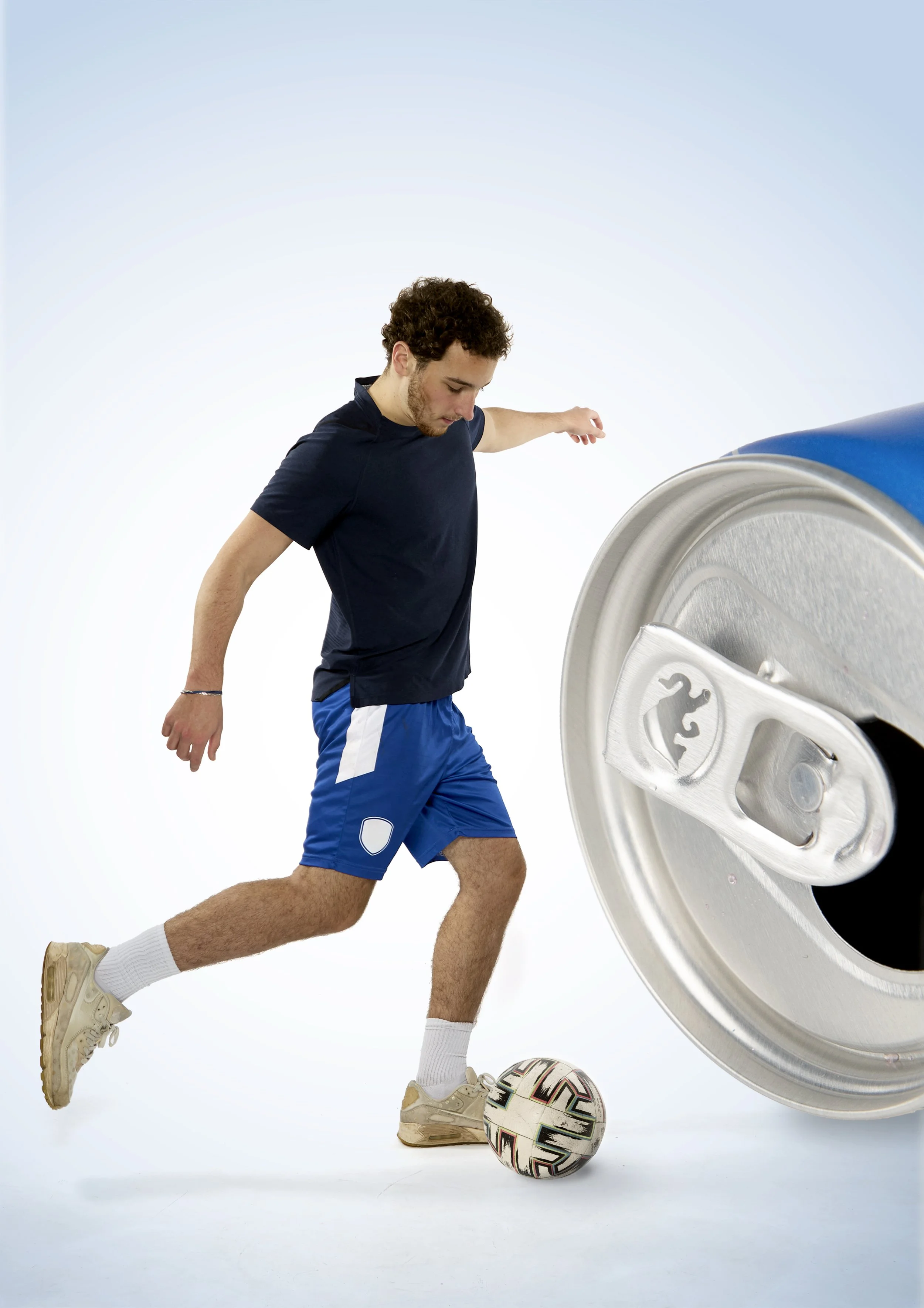 A young man playing soccer, wearing a navy blue t-shirt, blue shorts with white accents, white socks, and beige sneakers, is in mid-action to kick a soccer ball. There is an oversized aluminum soda can with a blue label in the background, tilted to o